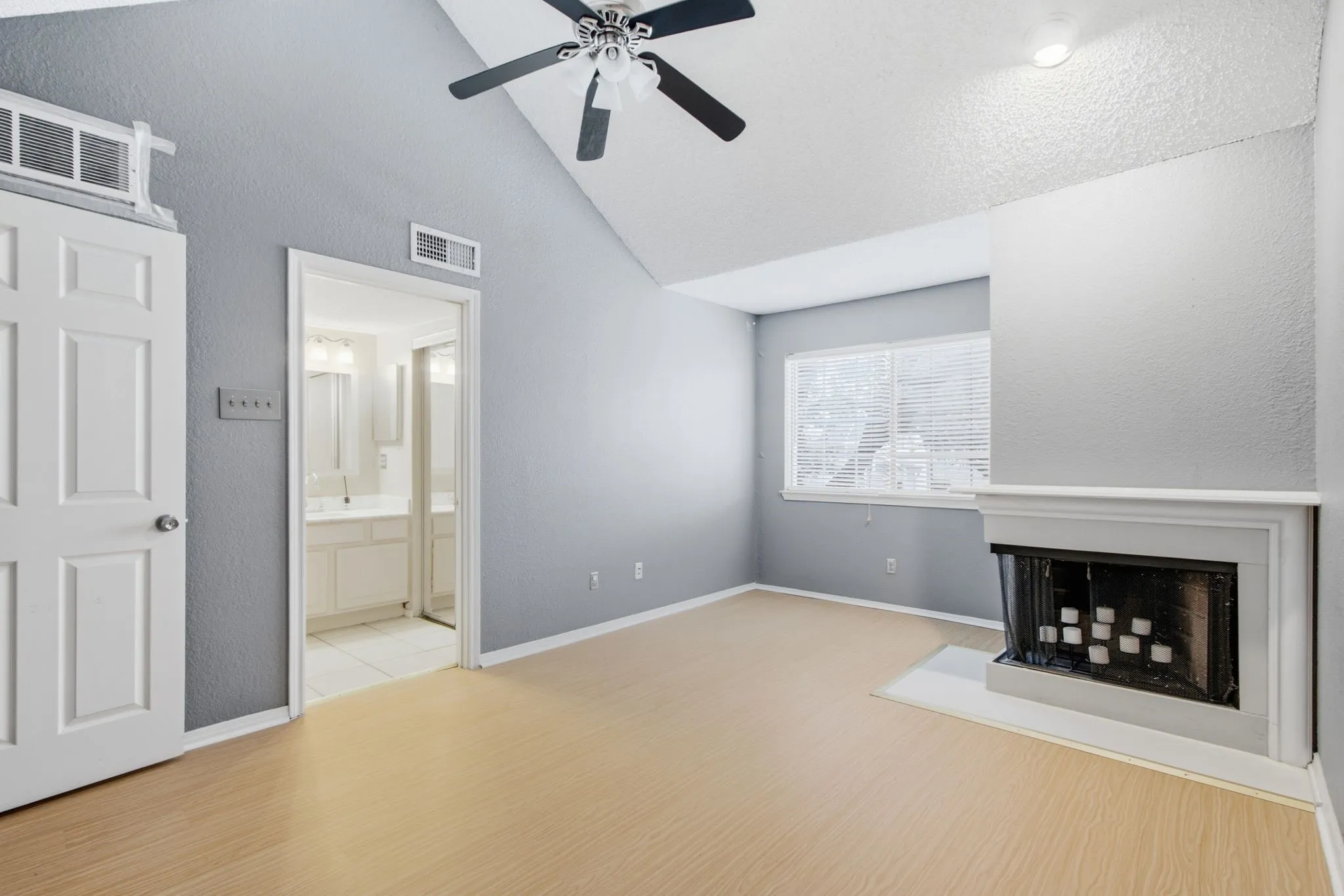 Vaulted ceiling room featuring wood-finish flooring, a ceiling fan, a window with blinds, and a white fireplace with a hearth