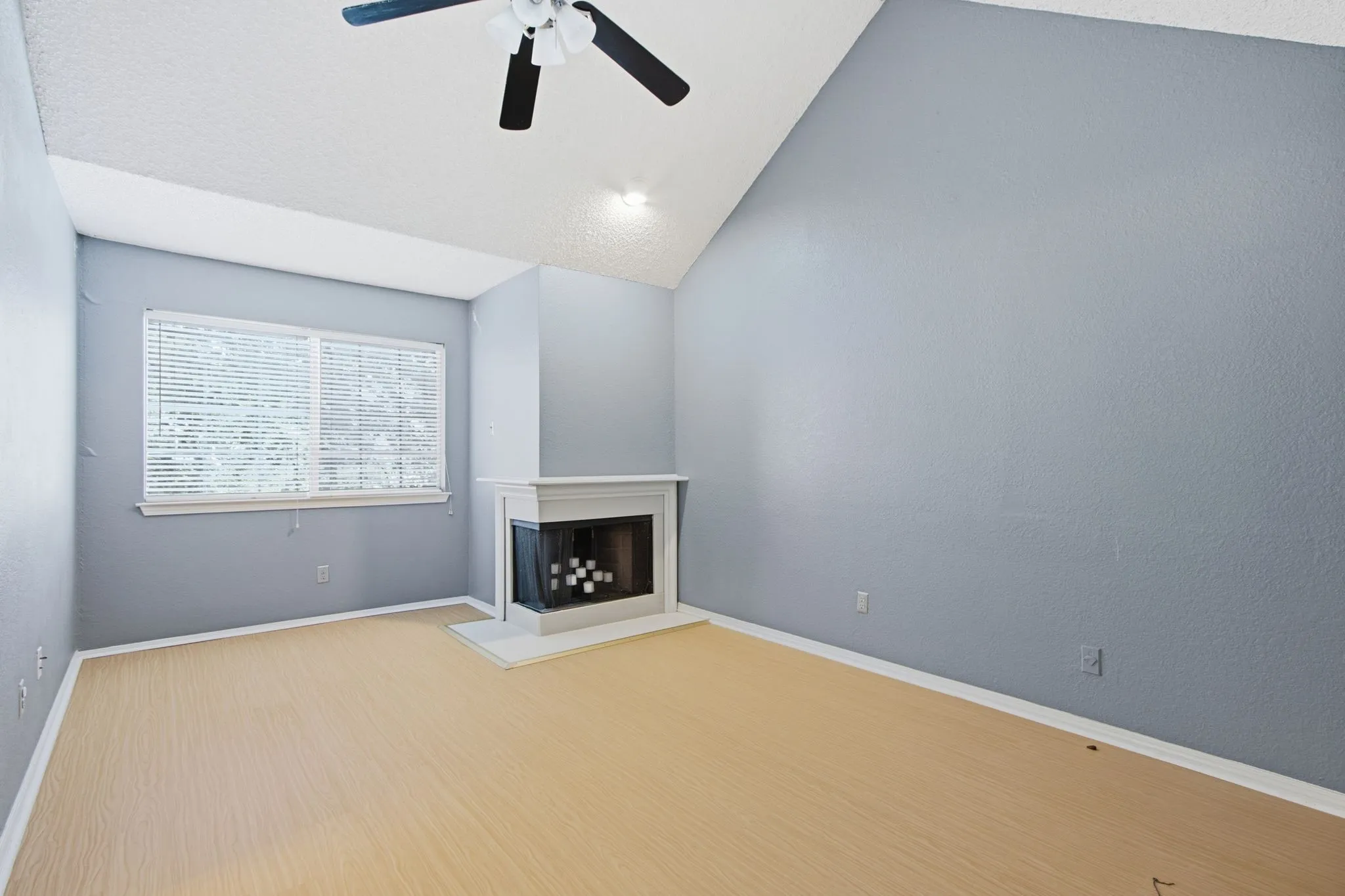 Vaulted ceiling room featuring a corner fireplace with white mantel and hearth, wood-finish flooring, a window with horizontal blinds, and a ceiling fan with integrated lighting