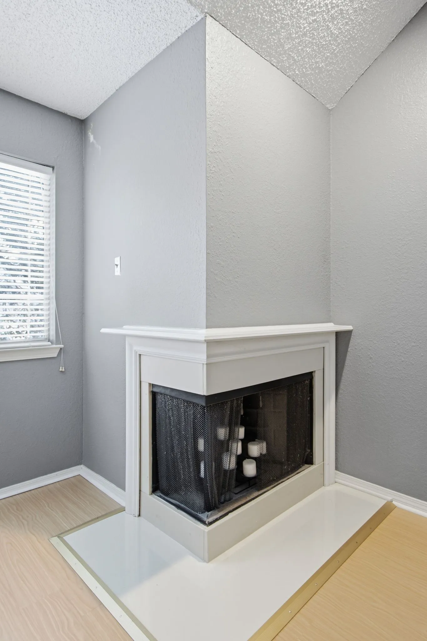 Corner fireplace with a white mantel and hearth, light wood-finish flooring, and a window with blinds