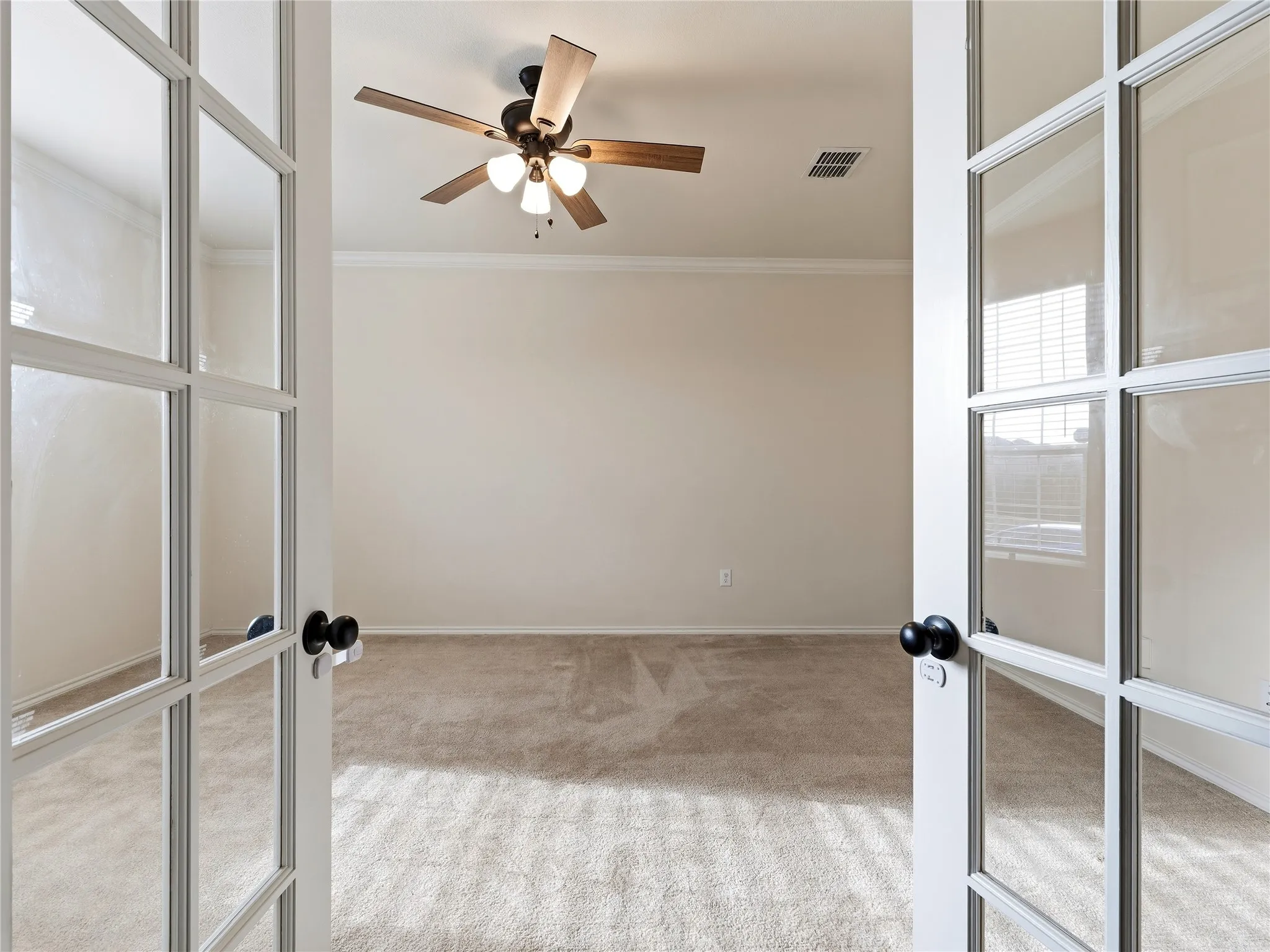 French doors with glass panels opening to an interior room with neutral carpet flooring, ceiling fan with light fixture, baseboard molding, and crown molding