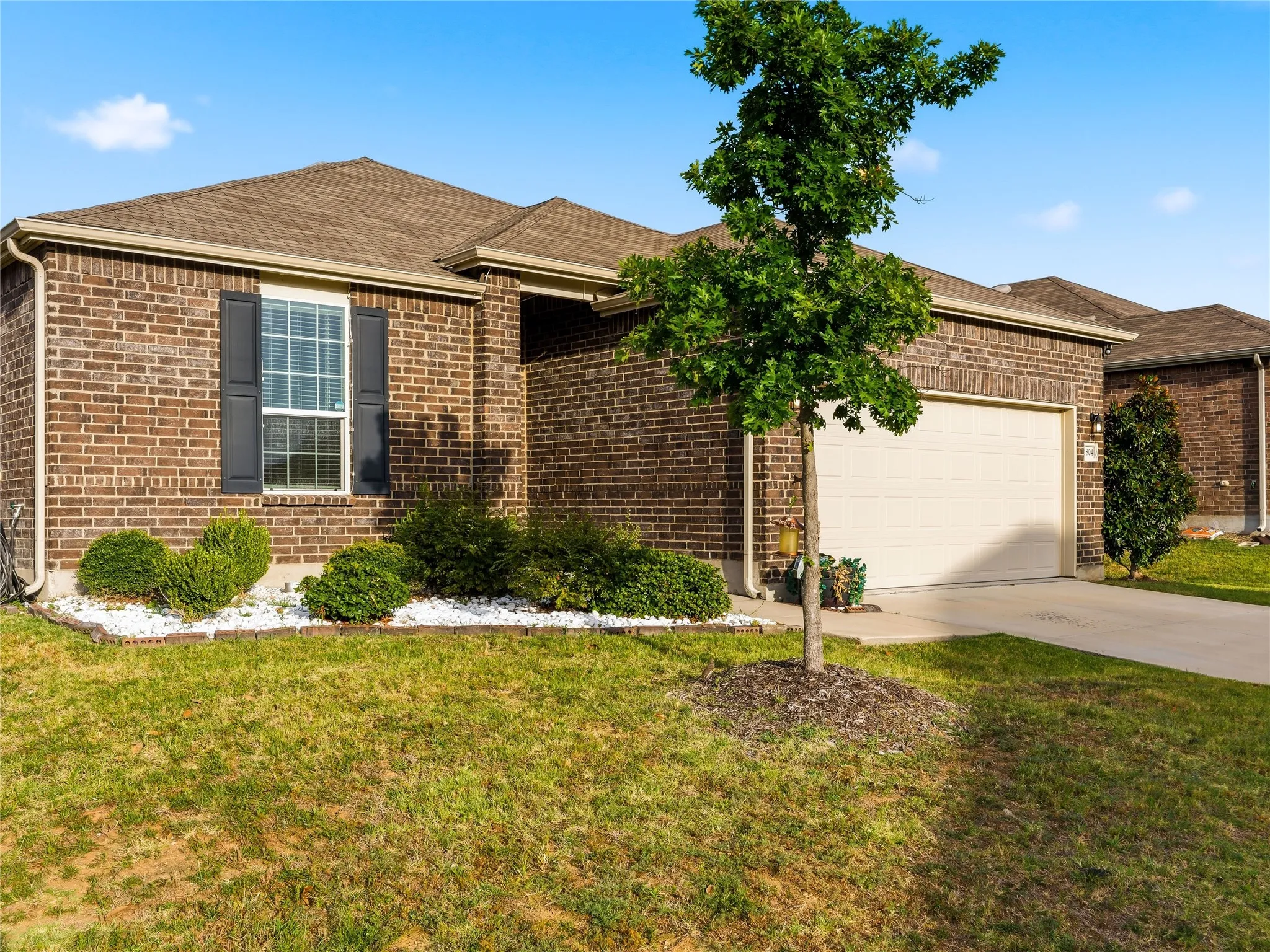 Brick exterior featuring a single window with dark shutters, an attached two-car garage, concrete driveway, and front landscaping with white decorative rocks and established shrubs