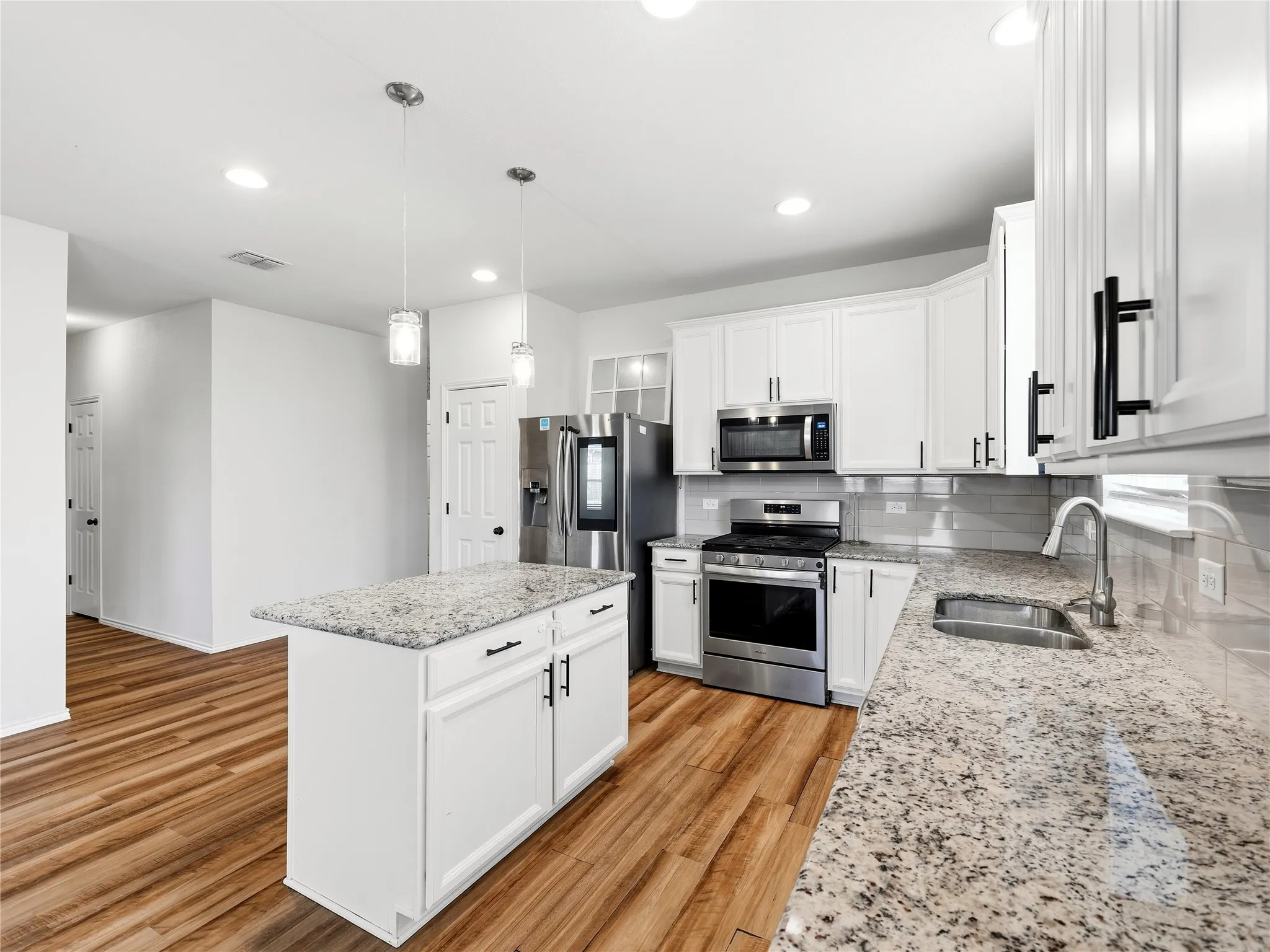 Kitchen featuring wood-finish flooring, white cabinetry with black hardware, stainless steel appliances, a tile backsplash, and a central island with a stone-finish countertop