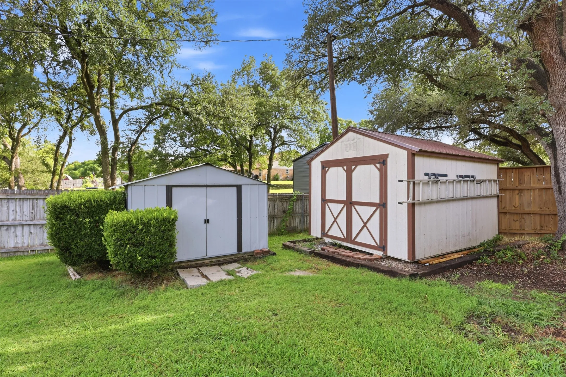 Expansive backyard featuring a metal storage shed and a wood-frame storage shed with double doors, surrounded by mature trees and a wood privacy fence