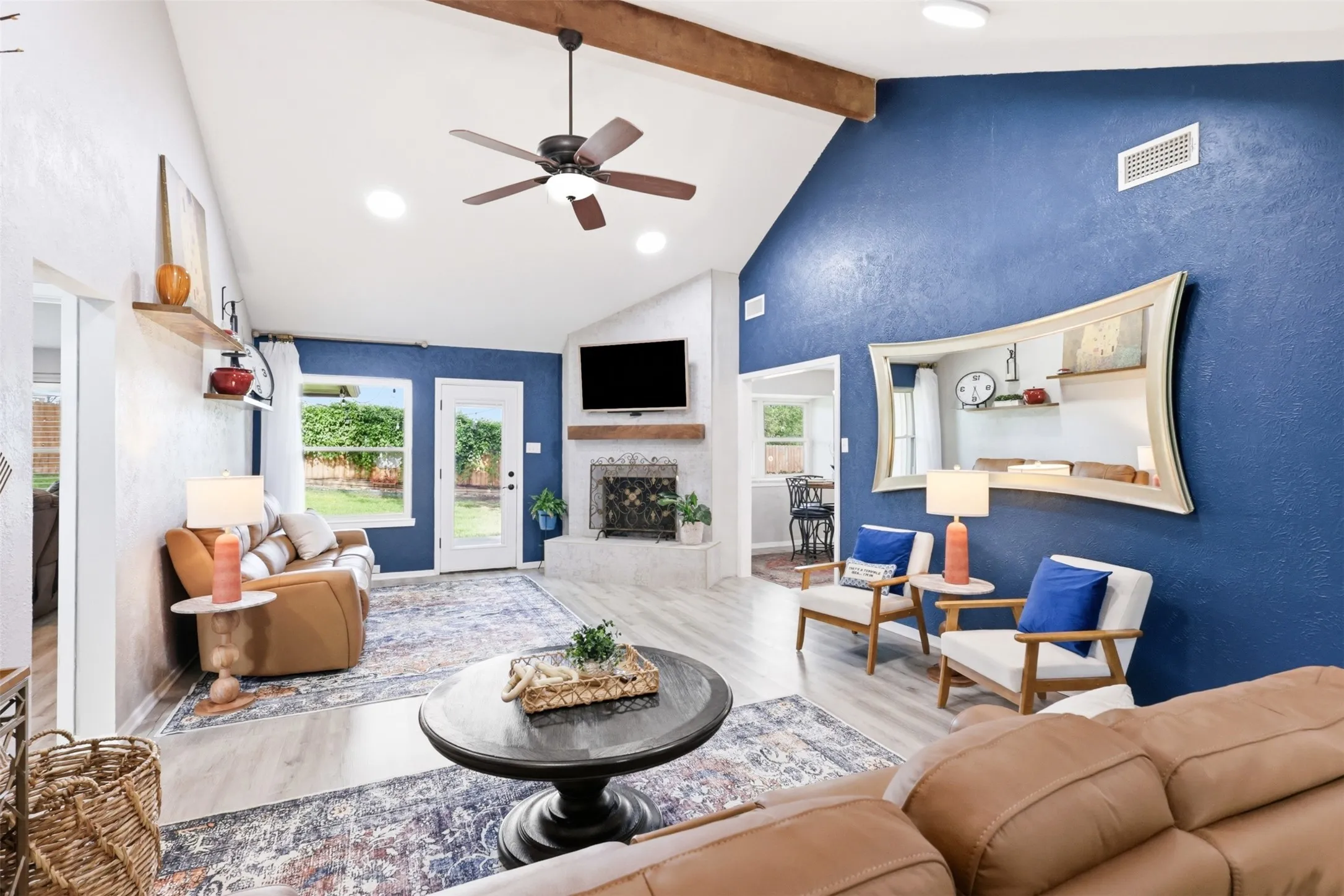 Living area featuring a vaulted ceiling with exposed beams, wood-finish flooring, a white brick fireplace with a wood mantle, a ceiling fan, and recessed lighting