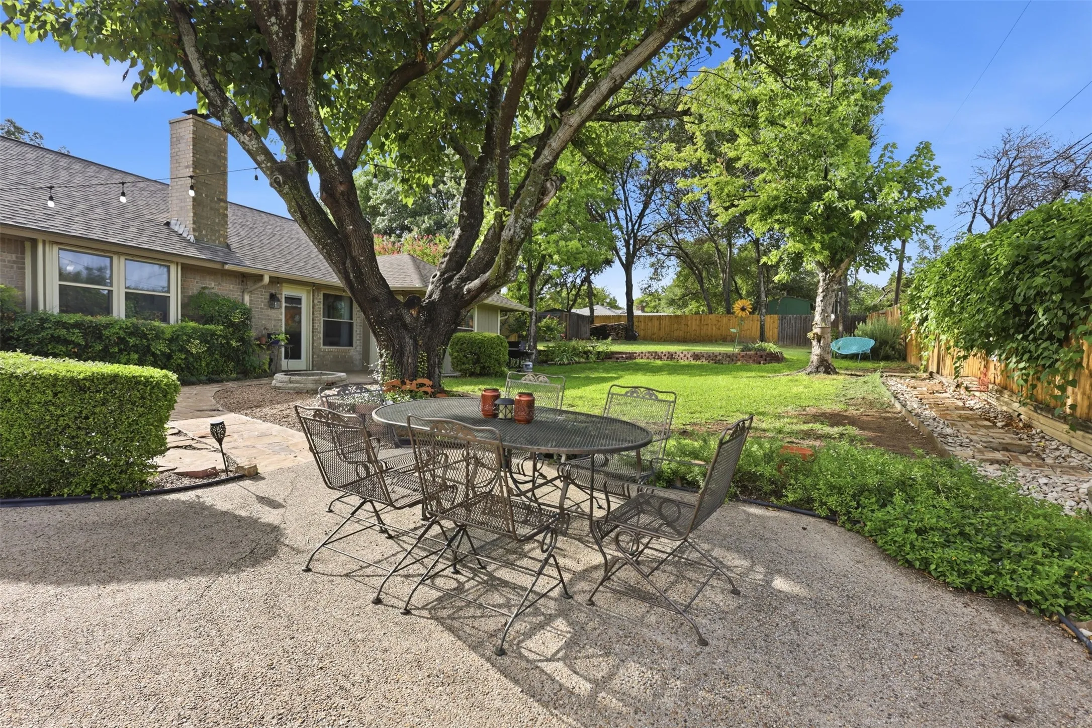 Spacious backyard featuring a large mature tree, a concrete patio, a stone pathway, a manicured lawn area, and a brick exterior with multiple windows