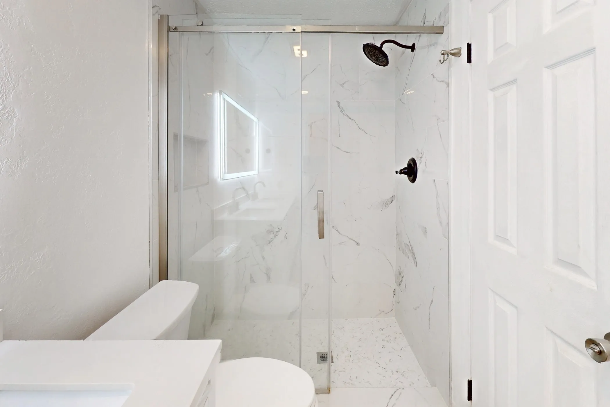 Contemporary bathroom featuring a frameless glass shower enclosure, white marbled wall tiles, black shower fixtures, a built-in shower niche, and a white toilet