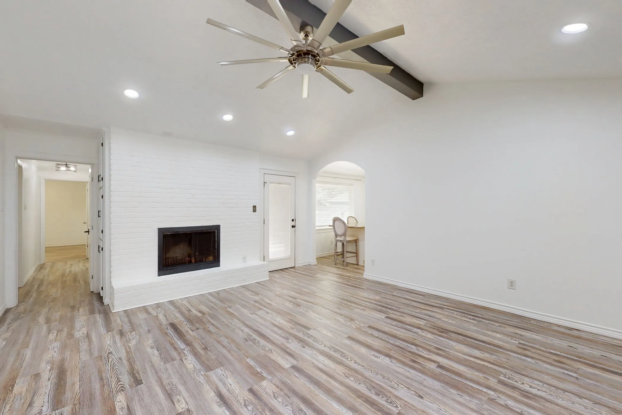 Spacious living area featuring wood-finish flooring, a white-painted brick fireplace, vaulted ceilings with a ceiling fan, recessed lighting, and an arched doorway leading to an additional space