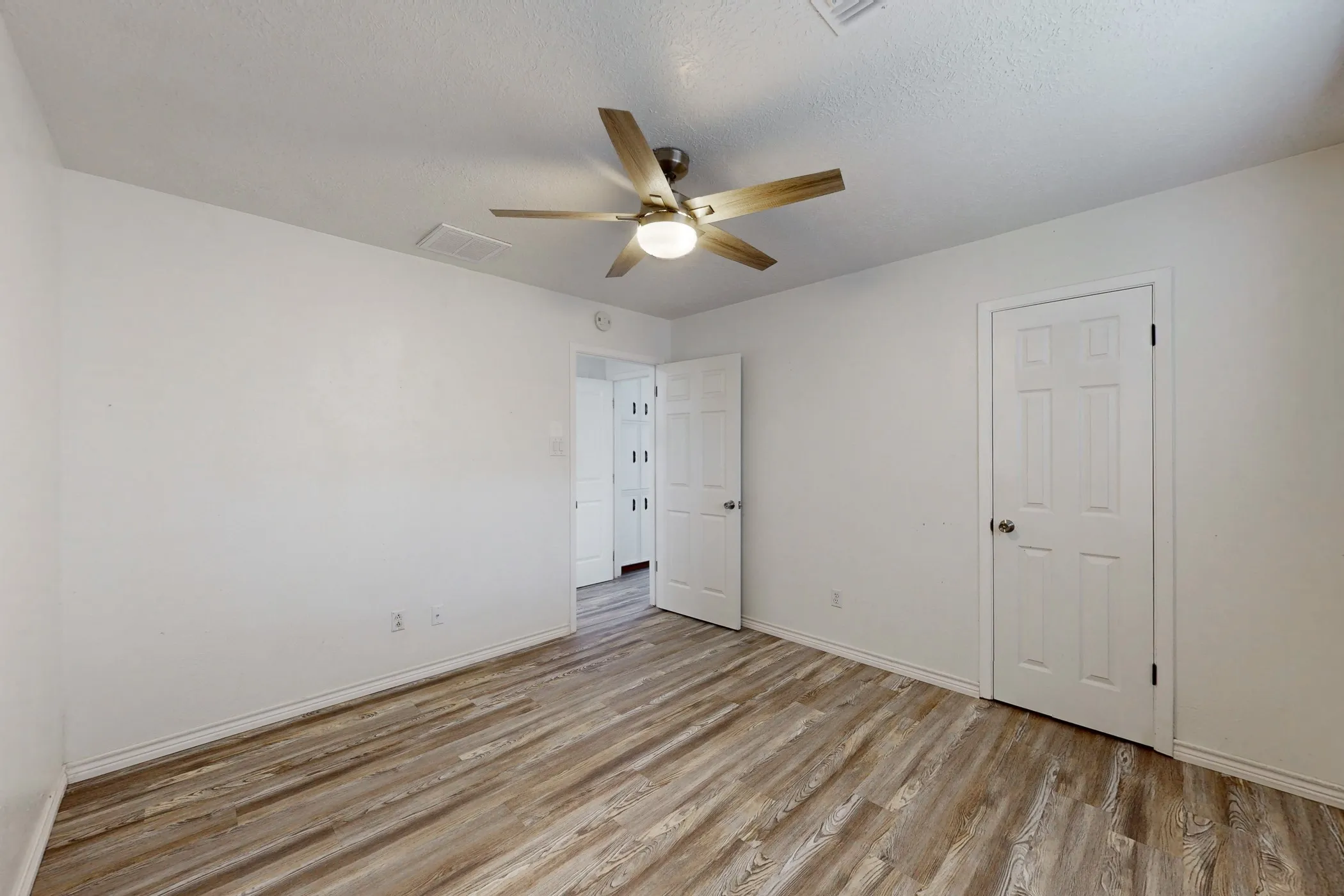 Spacious room with wood-finish flooring, crisp white walls, a ceiling fan with integrated lighting, and white paneled doors