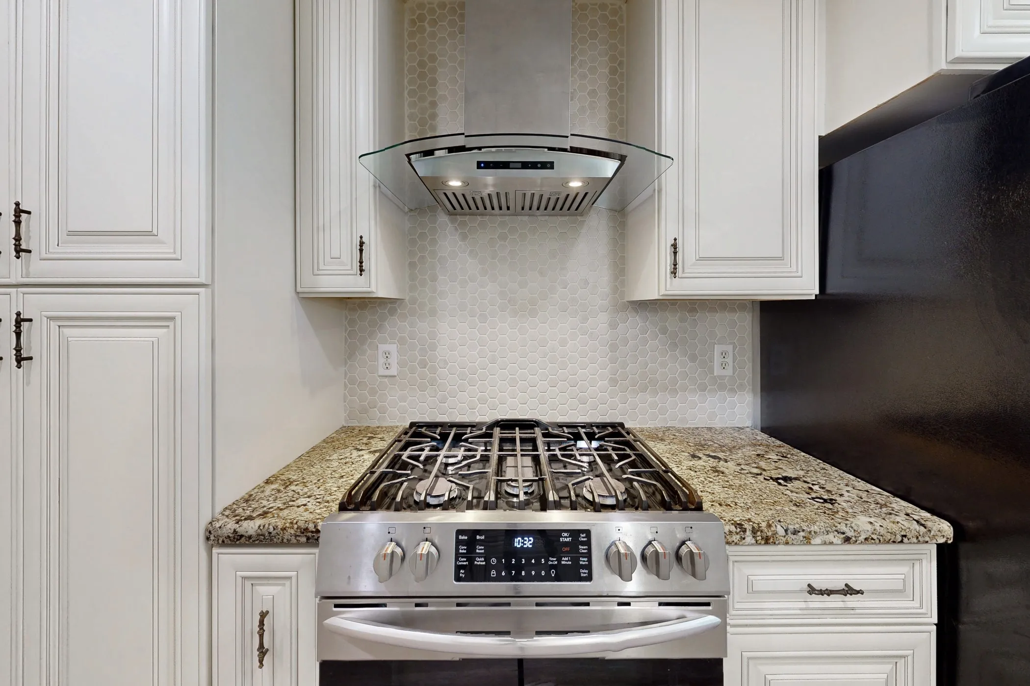 Stainless steel gas range with coordinating vent hood, cream-toned cabinetry, honeycomb tile backsplash, and granite countertops