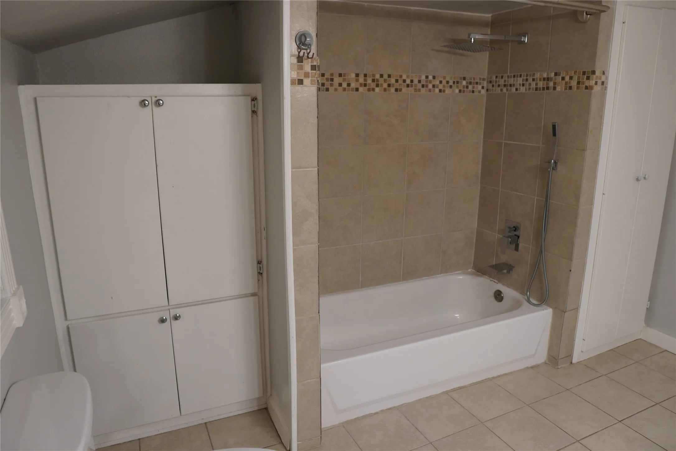 Bathtub with tan tile surround, decorative mosaic accent tile, and a rain shower head