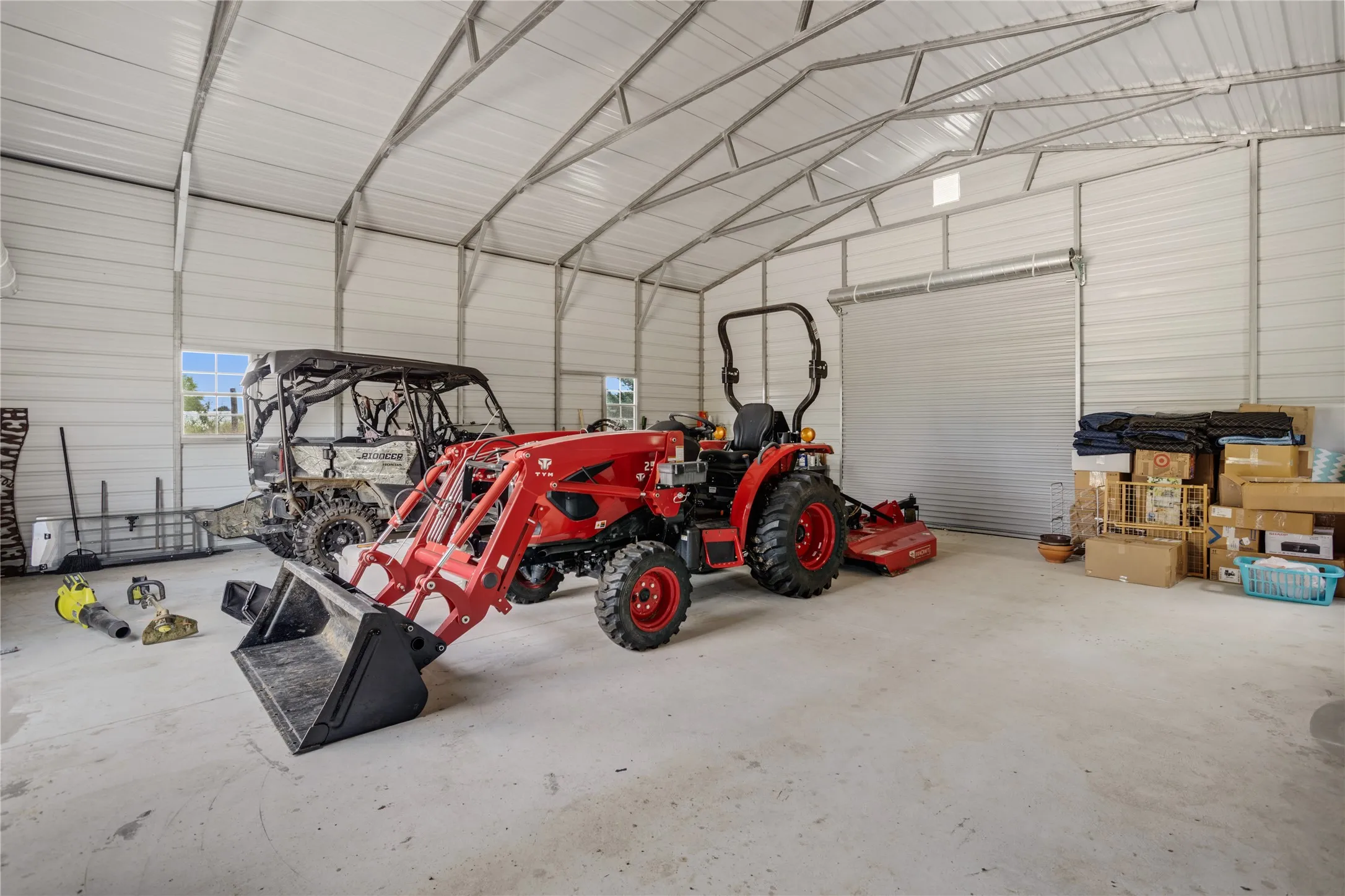 Spacious metal building featuring a concrete floor, high vaulted ceilings with exposed metal trusses, multiple windows for natural light, and a large roll-up door