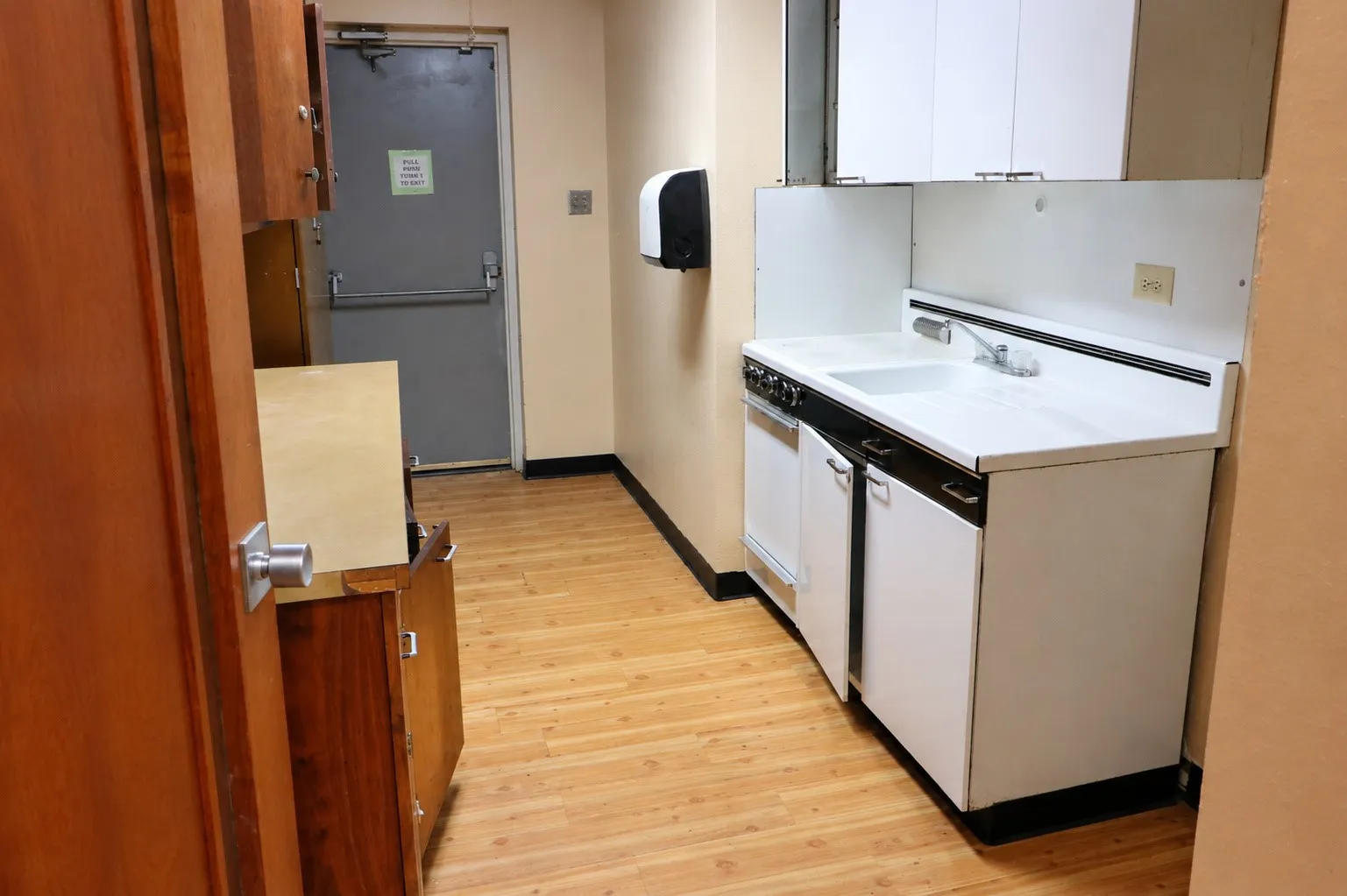 Compact kitchenette featuring a white countertop with integrated sink, white upper and lower cabinetry, and wood-finish flooring