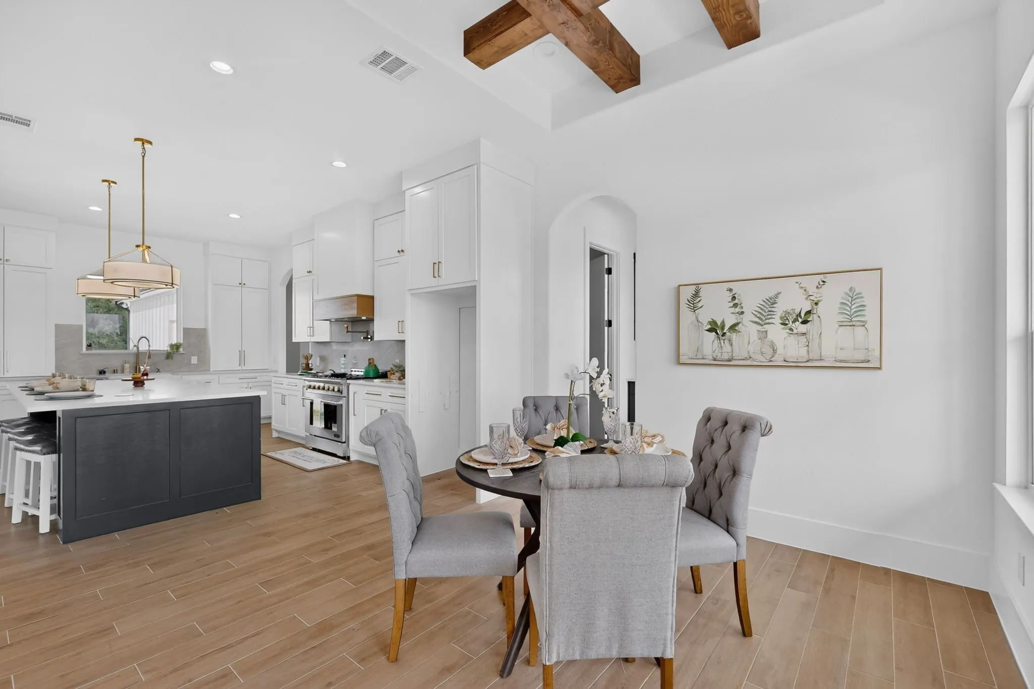 Bright open-concept kitchen and dining area featuring wood-finish flooring, a white cabinet kitchen, a dark-toned kitchen island with seating, pendant lighting, and exposed ceiling beams