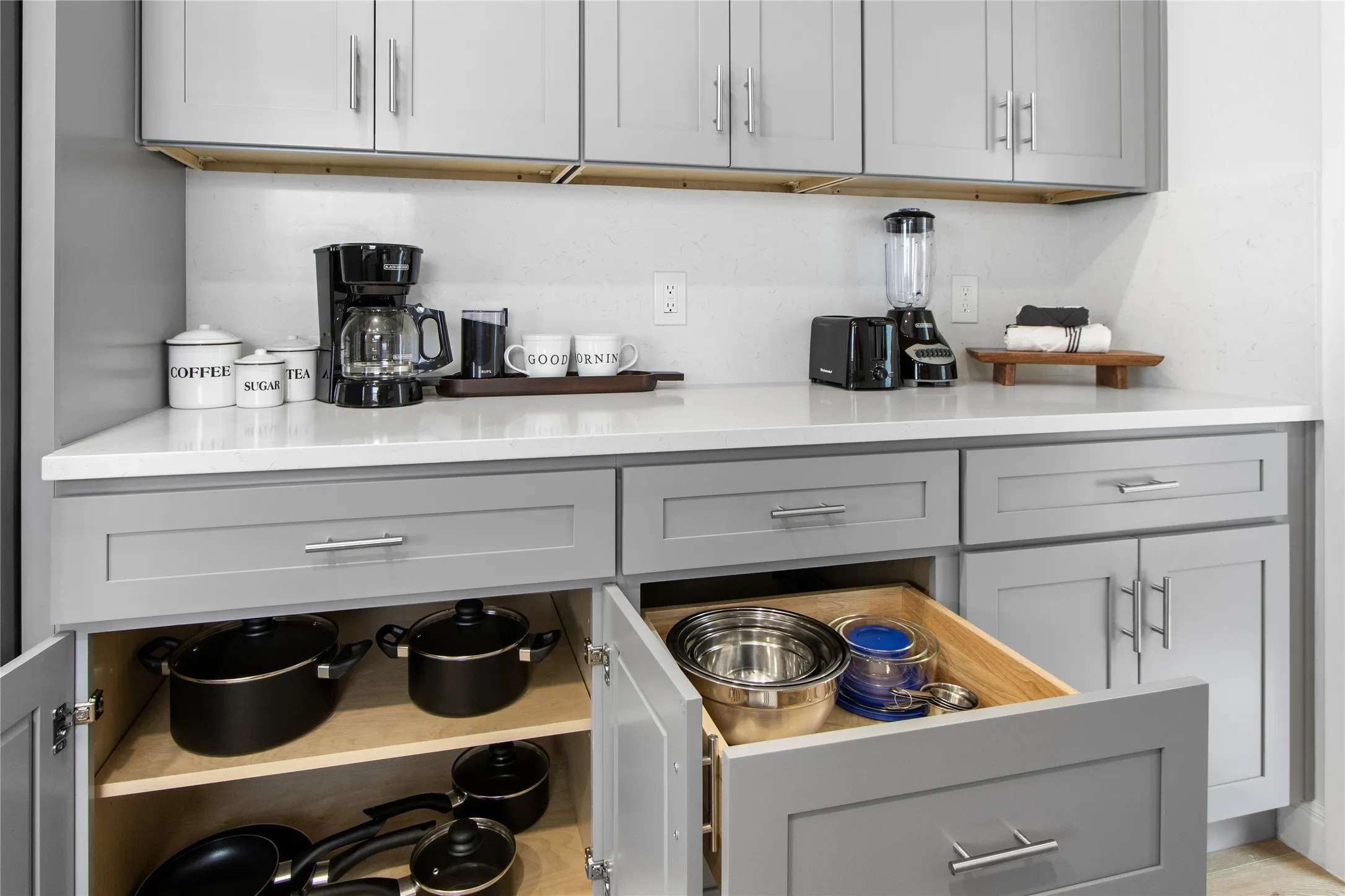 Gray shaker-style cabinetry with chrome hardware, white solid surface countertops, and light-toned backsplash