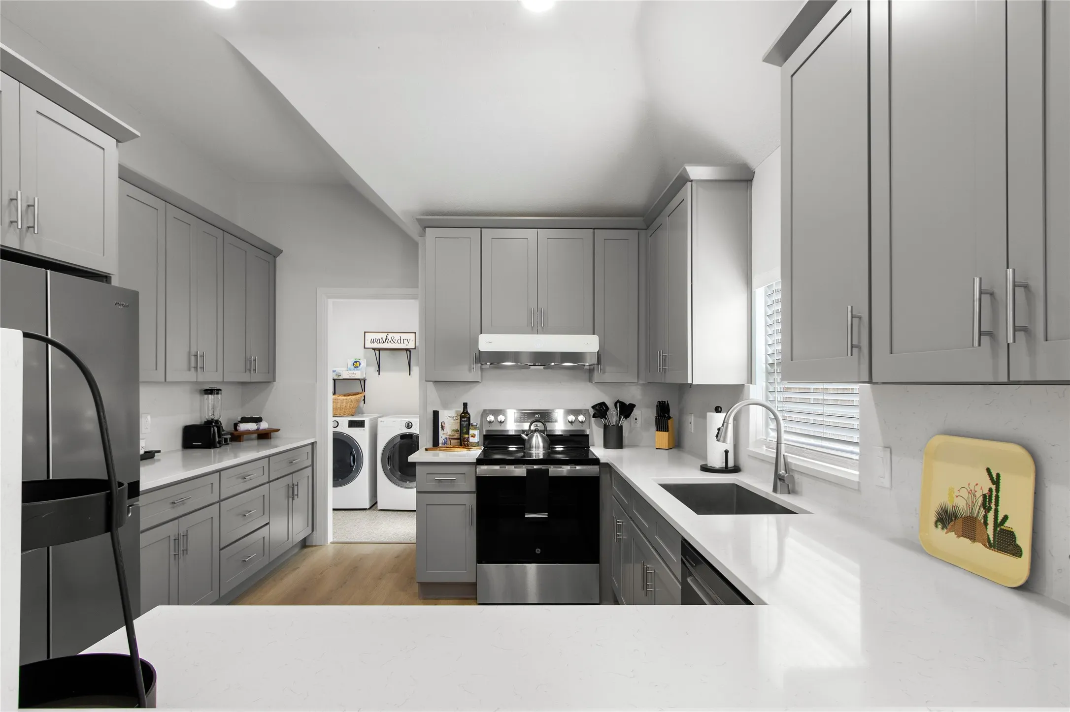 Modern kitchen featuring gray shaker-style cabinetry, white countertops, wood-finish flooring, a stainless steel range, and a laundry area