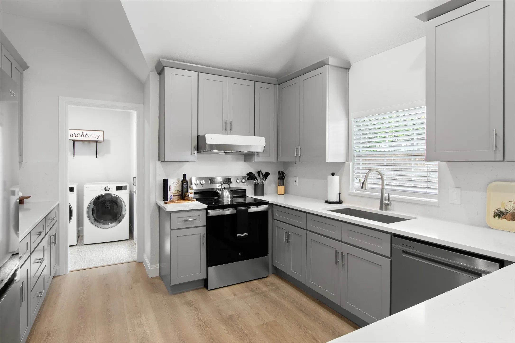 Contemporary kitchen featuring light gray shaker cabinetry, white countertops, wood-finish flooring, and stainless steel appliances including a range and dishwasher