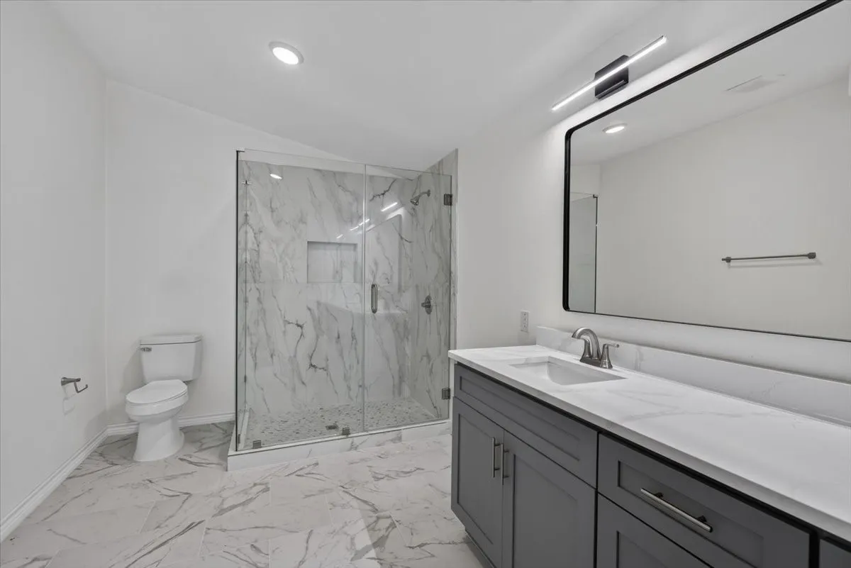 Bathroom featuring a large vanity with a white countertop and double sinks, a frameless glass shower enclosure with marble-patterned tile, and recessed lighting