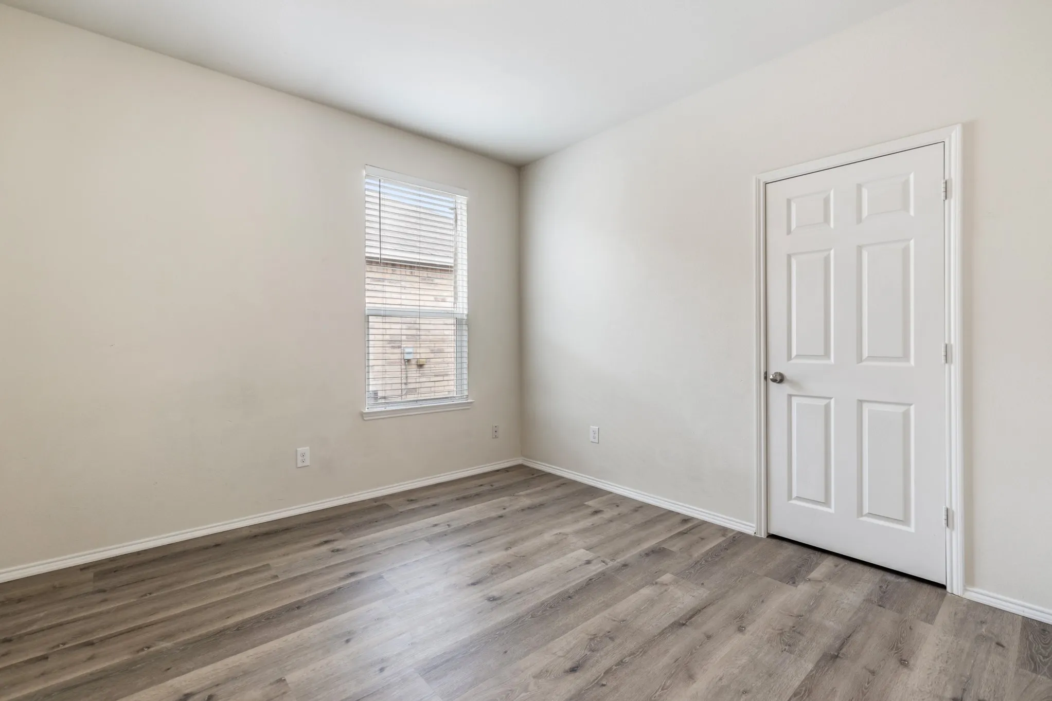 Room featuring light-toned walls, wood-finish flooring, a paneled door, and a window with blinds