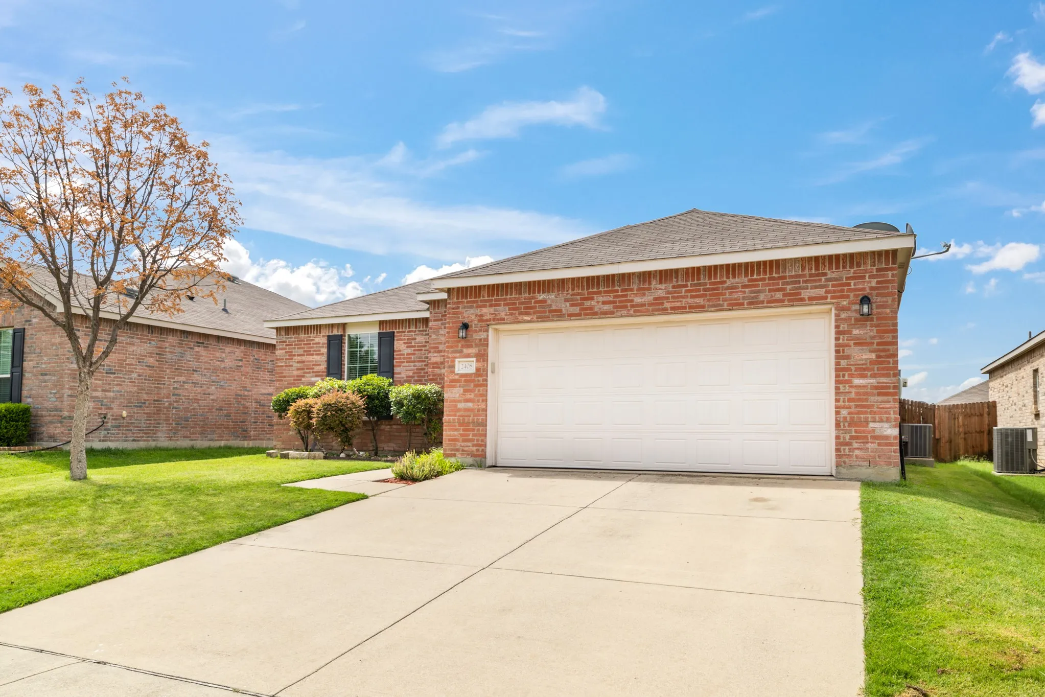 Brick exterior with an attached garage, concrete driveway, and established front landscaping