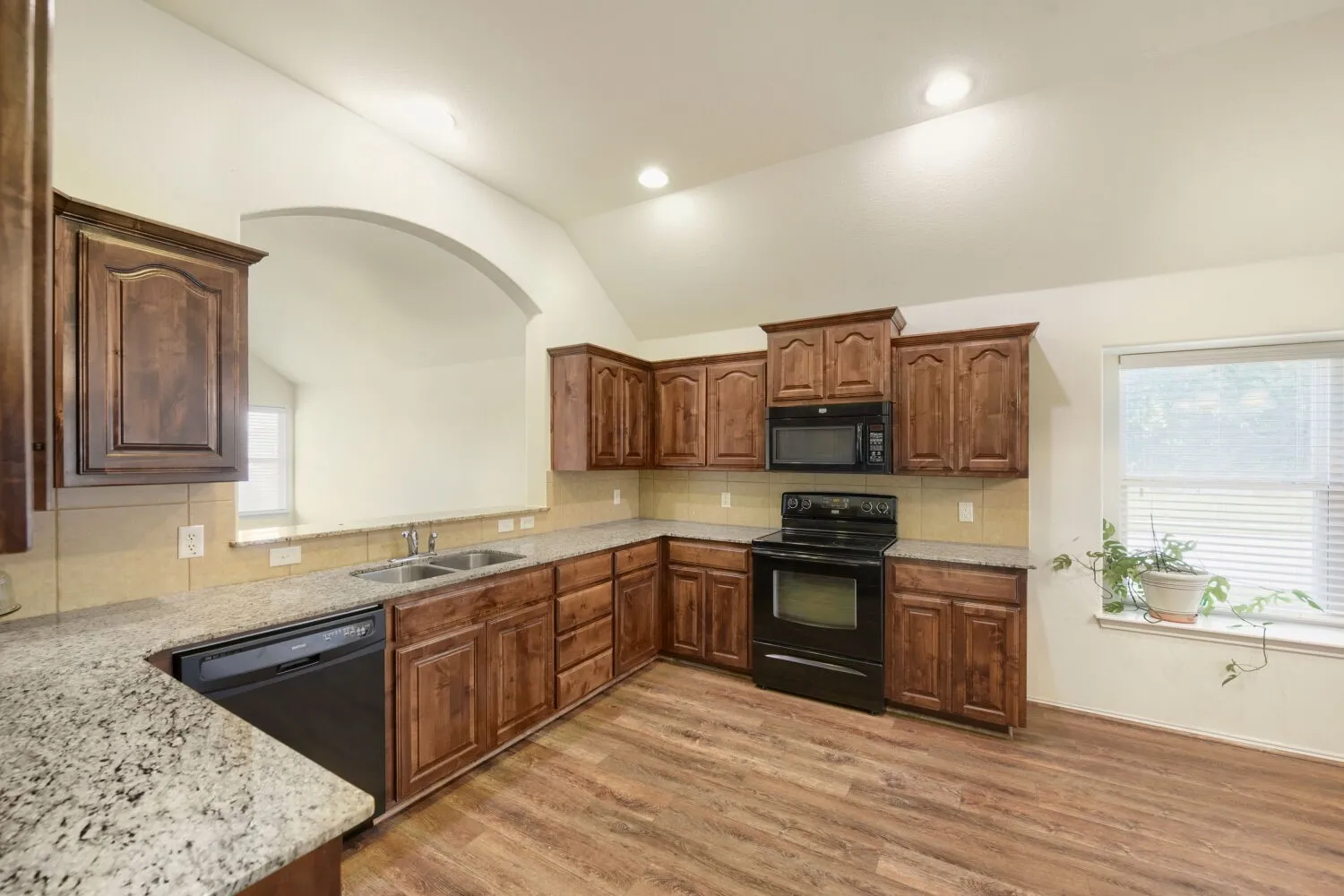 Kitchen featuring wood-finish flooring, granite-style countertops, wood cabinetry, built-in black appliances, and a pass-through arched opening