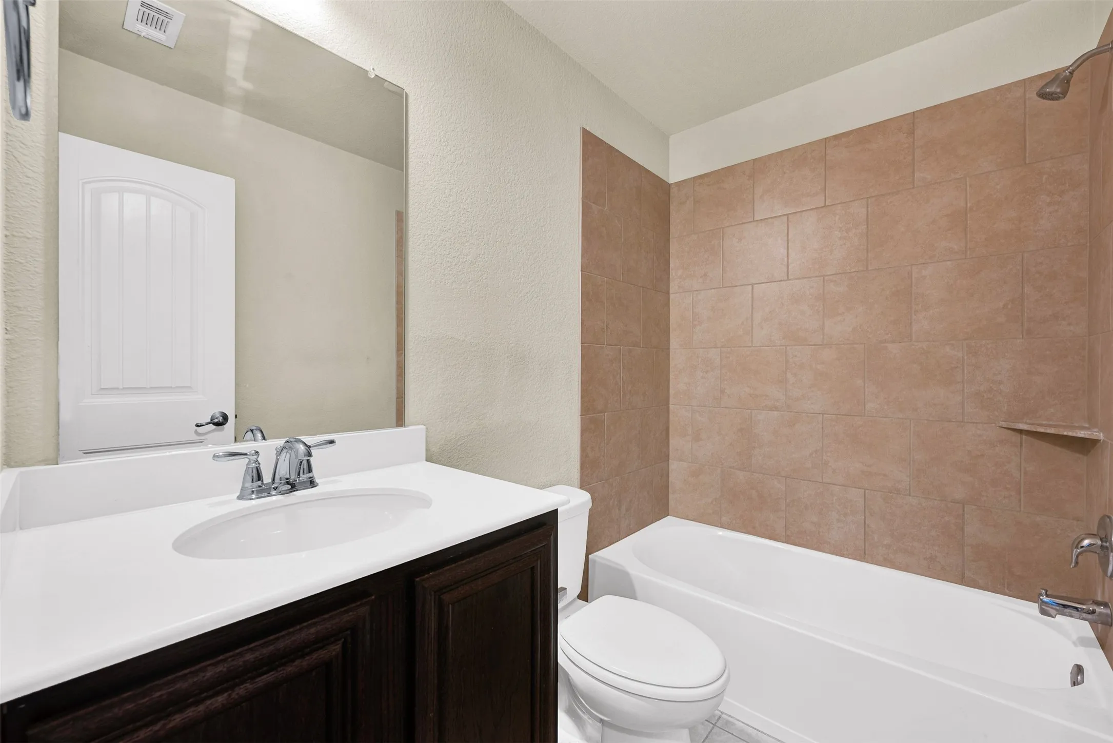 Bathroom featuring a white vanity with an integrated sink and chrome fixtures, dark wood-finish cabinetry, a full-width mirror, and an adjacent tiled shower with a built-in shelf