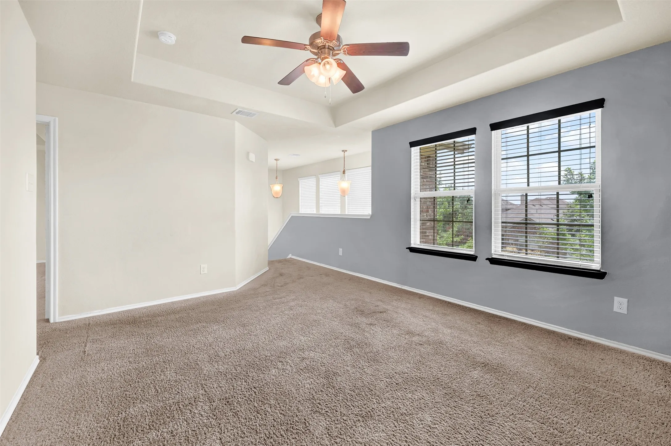 Spacious room with tray ceiling, ceiling fan, carpeted flooring, and a pair of windows featuring blinds