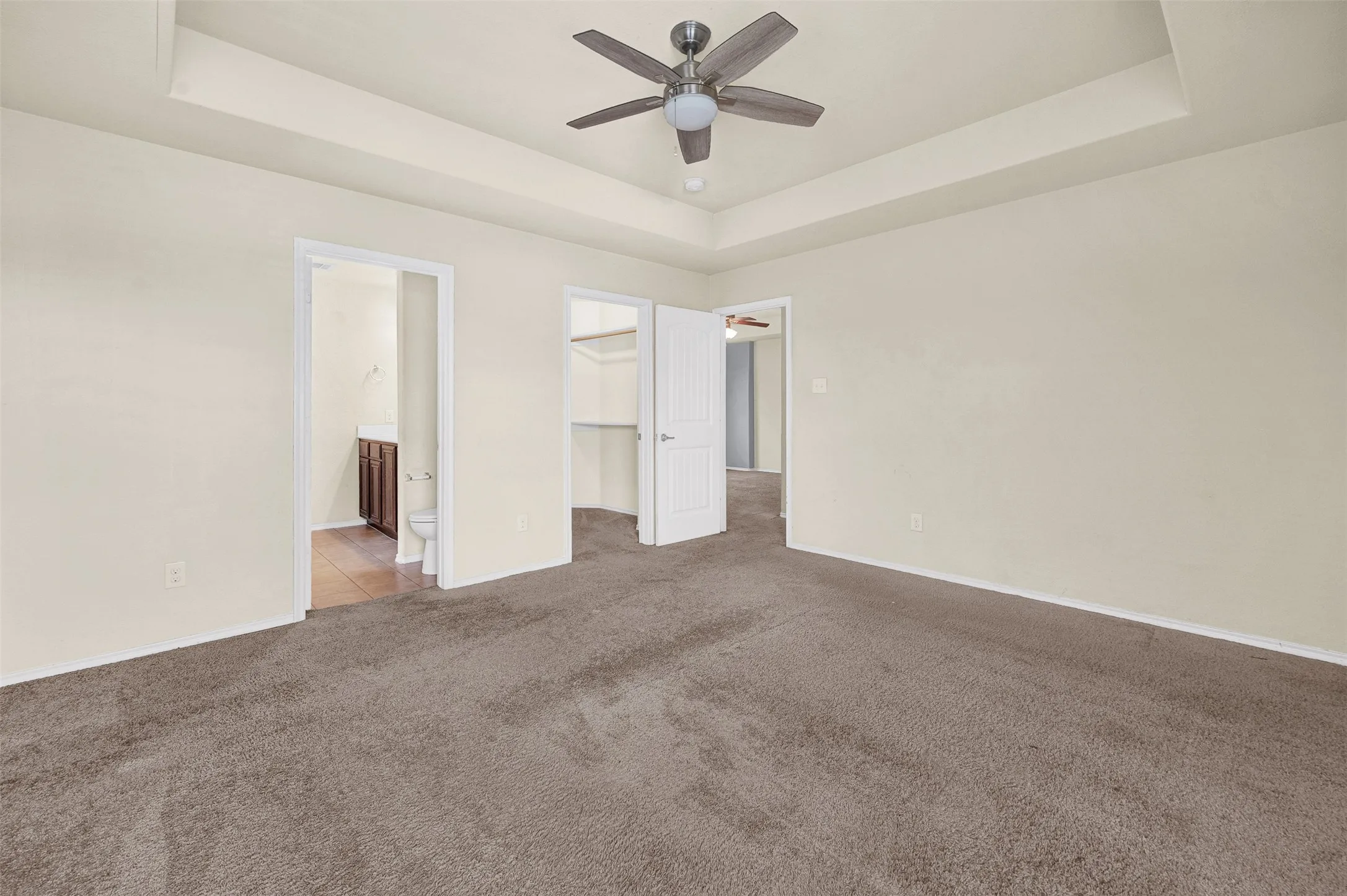 Carpeted bedroom featuring a tray ceiling with a ceiling fan, light-colored walls, and two doorways revealing a bathroom with tile flooring and a vanity, and a walk-in closet