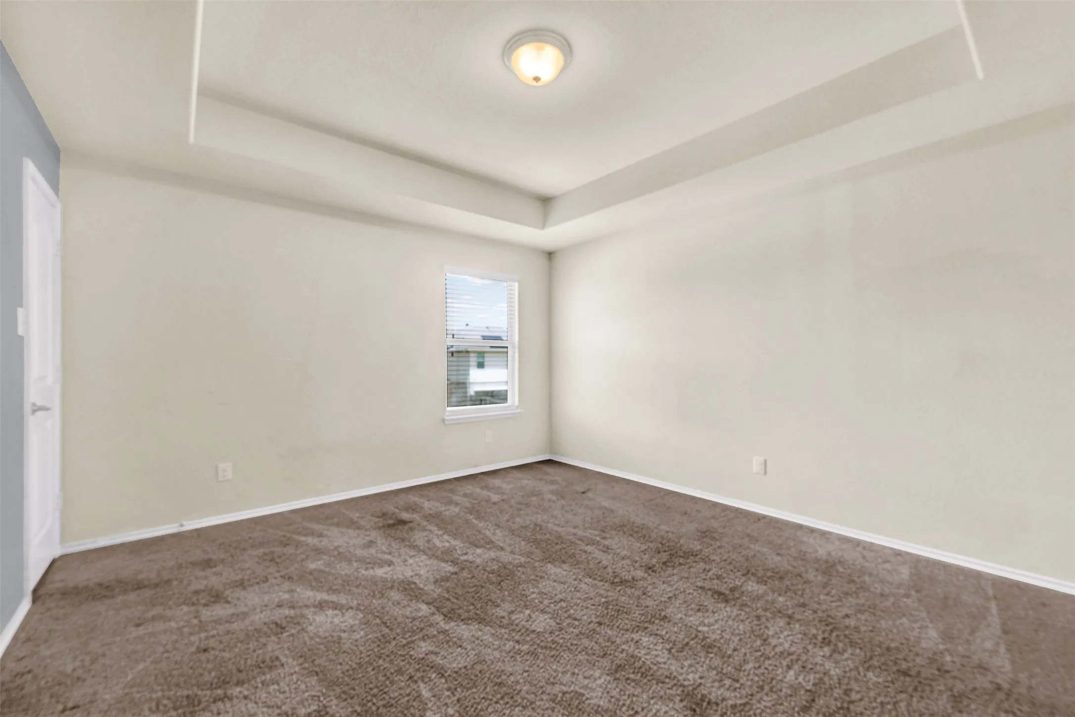 Spacious room featuring tray ceiling, flush mount lighting, single window with blinds, and brown carpet flooring