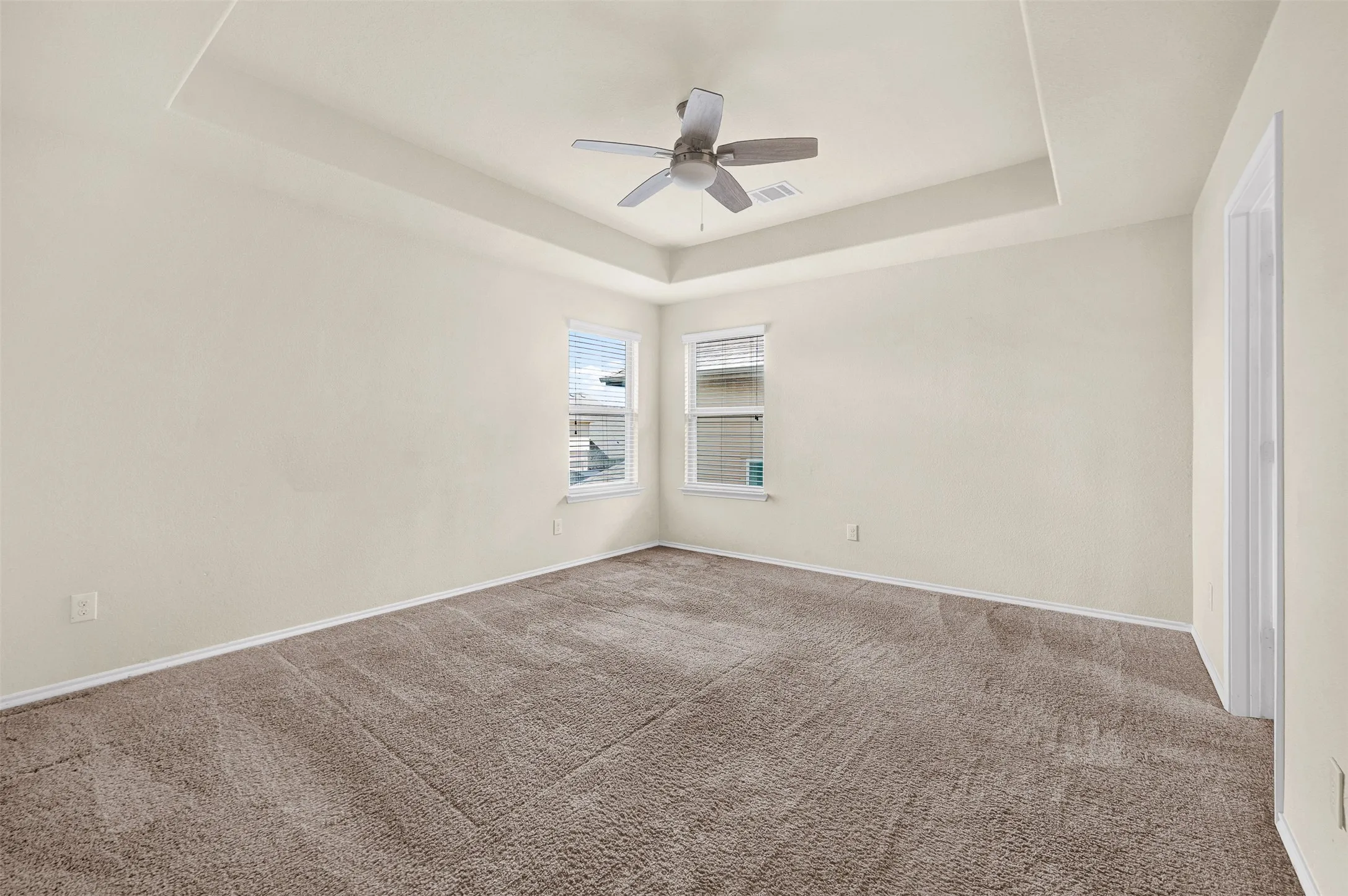 Spacious room featuring a tray ceiling, ceiling fan, two windows with blinds, and neutral carpeting