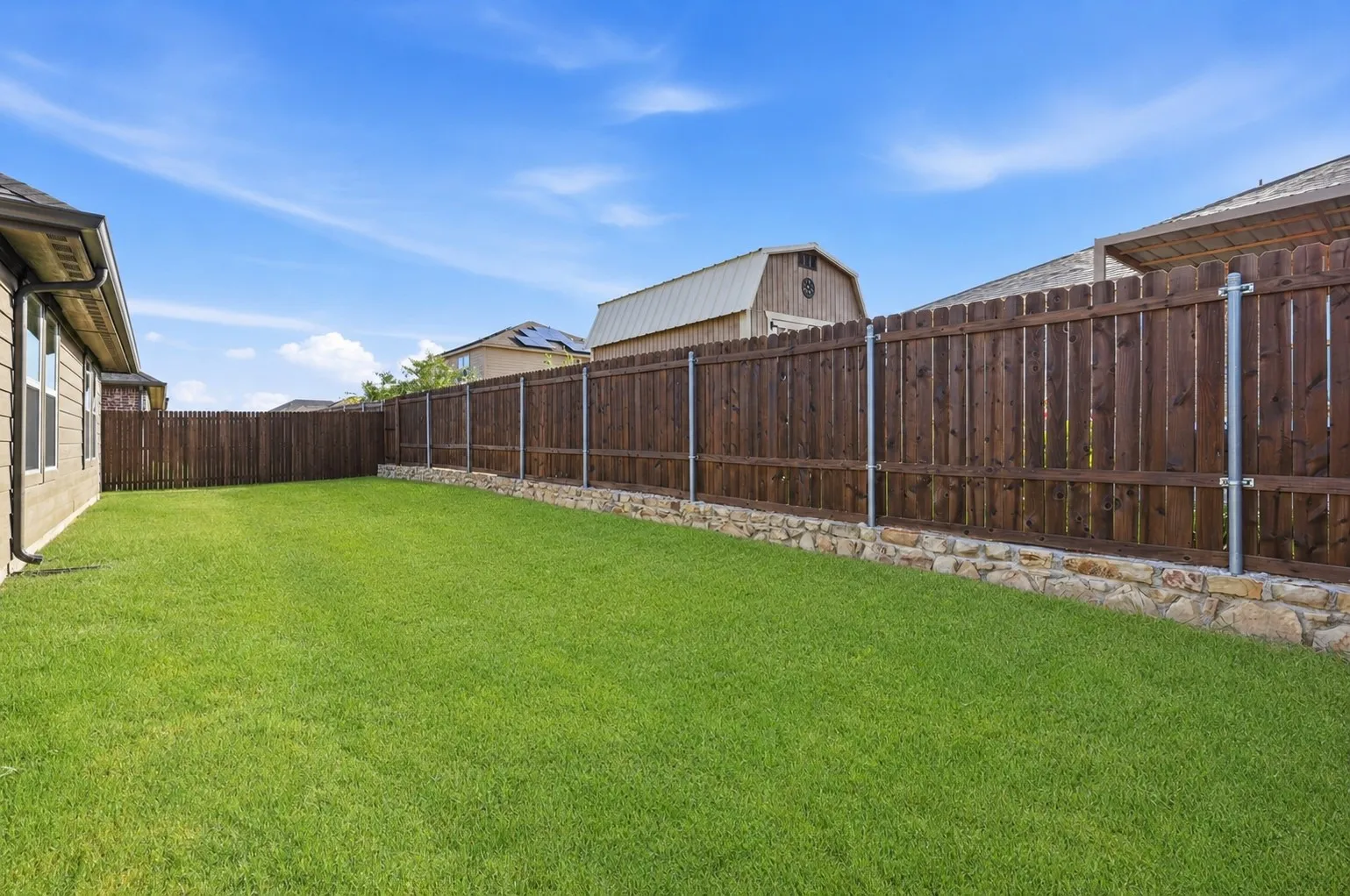 Expansive lawn area featuring a tall privacy fence with metal posts, a stone retaining wall, and a beige exterior wall with a gutter system
