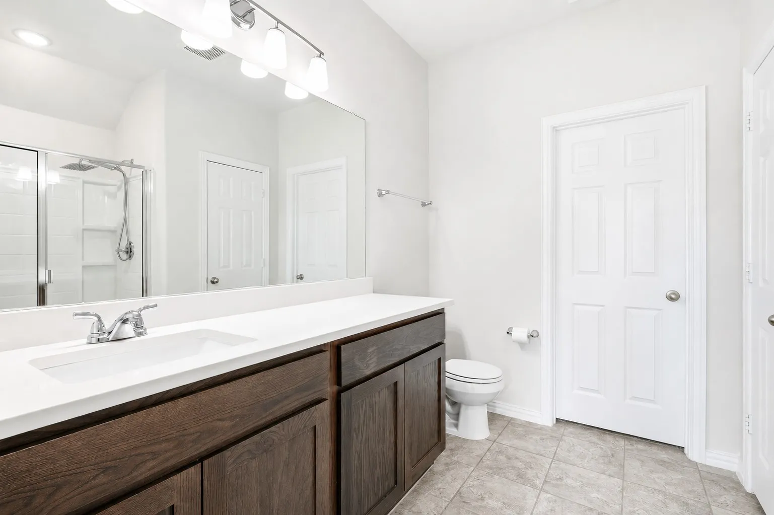 Bathroom featuring a wood-finish double vanity with a white countertop and integrated sinks, a mirror with a five-bulb light fixture, a tiled shower with a glass enclosure, and gray floor tiling