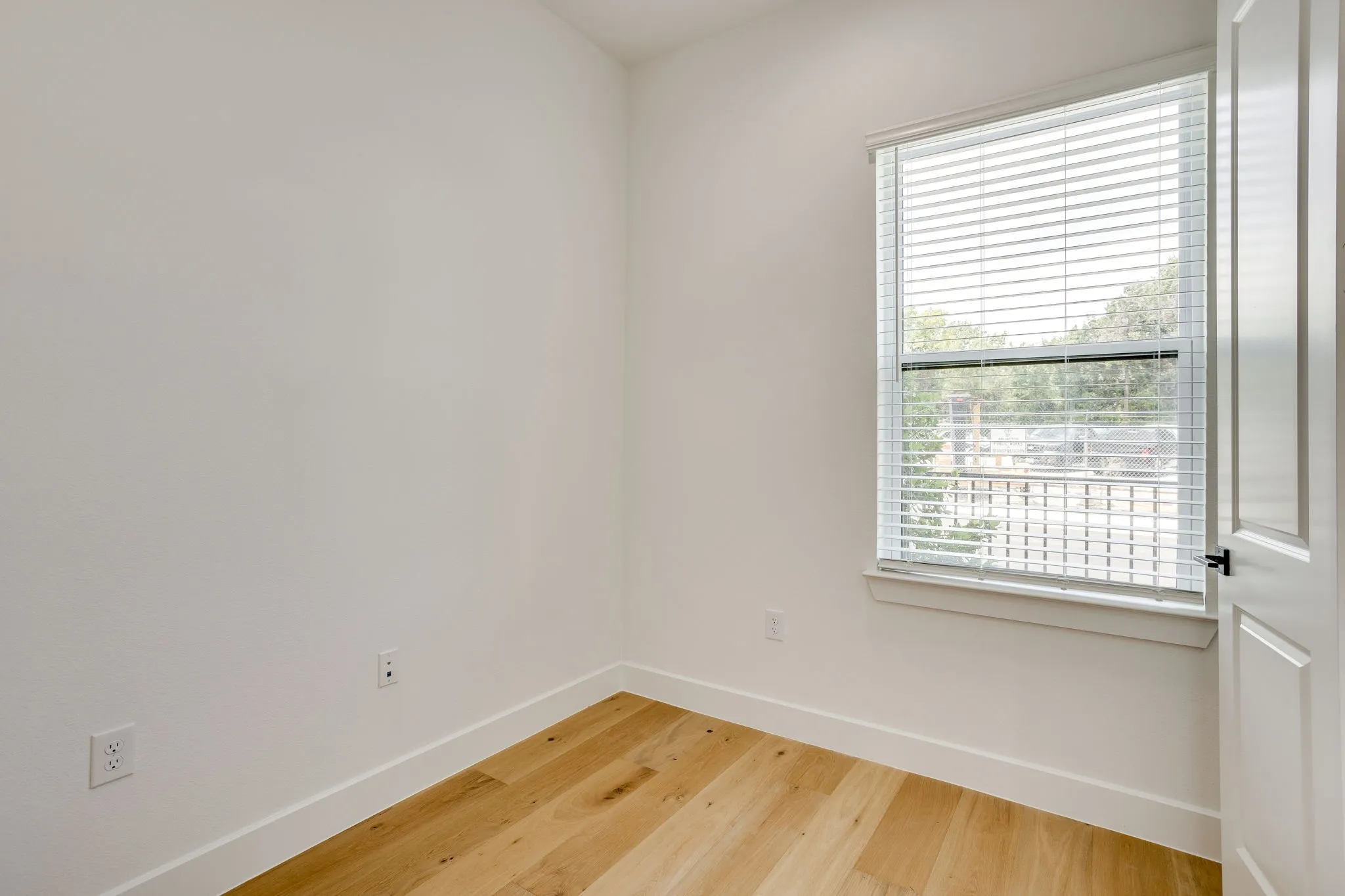 Bright room featuring a large window with horizontal blinds, light wood-finish flooring, white baseboards, and a white paneled door