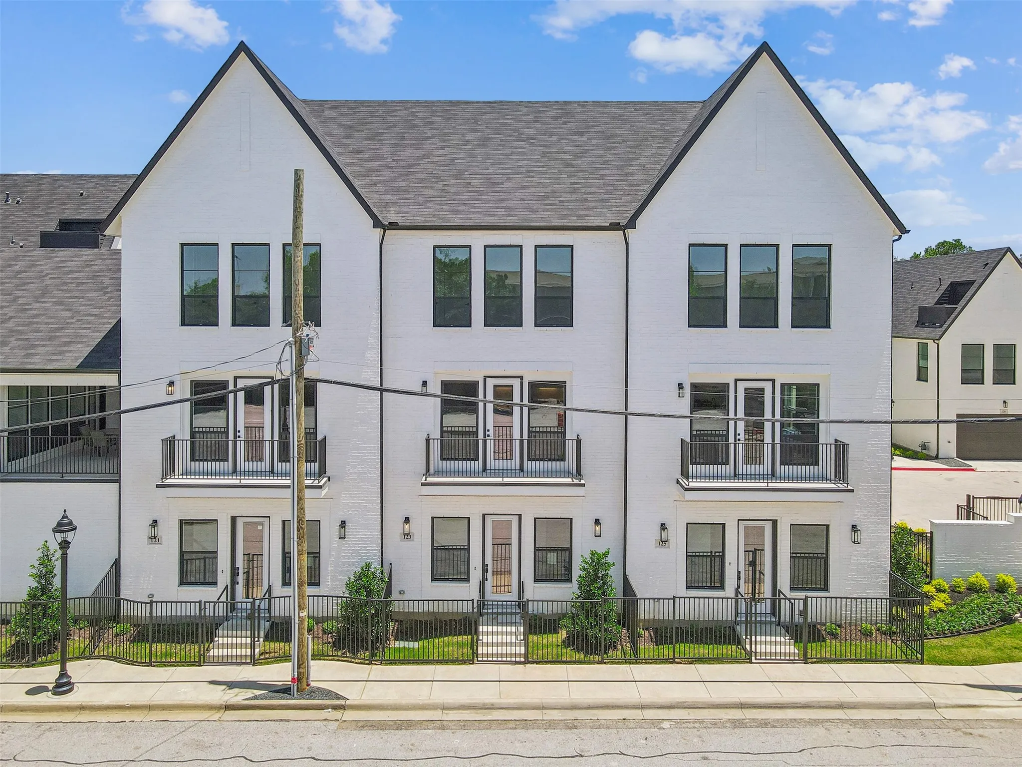 Contemporary townhome facade featuring white brick, black-framed windows, steep gables with dark shingle roofing, and individual iron-fenced front patios