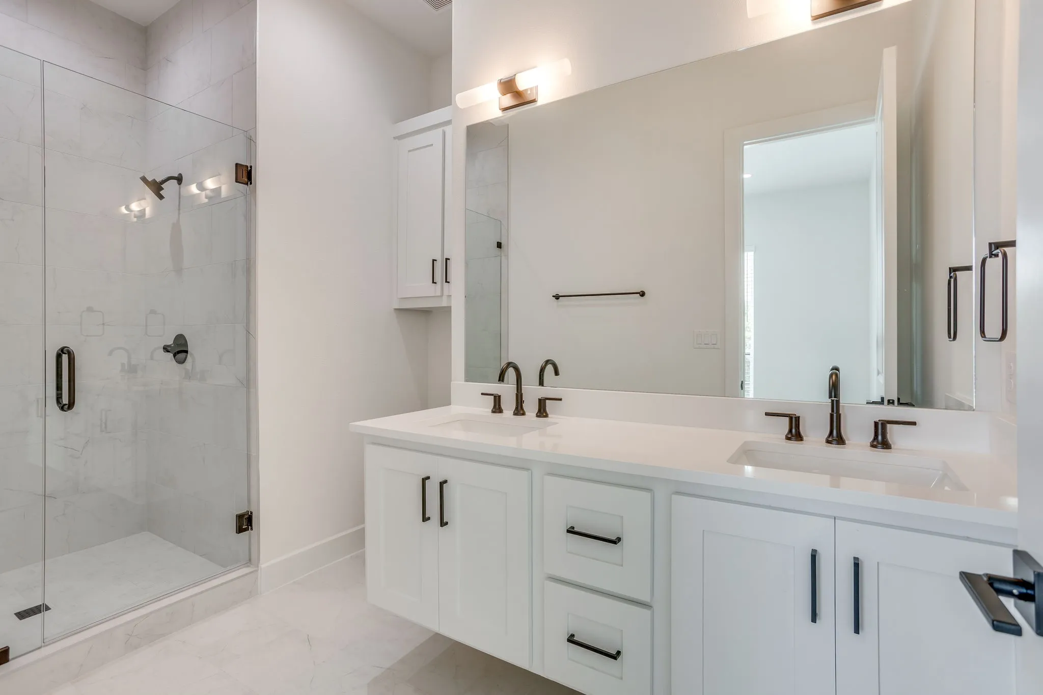 Contemporary bathroom featuring a spacious glass-enclosed shower with a multi-light fixture, dual vanity with white cabinetry and integrated sinks, a large wall-mounted mirror, and dark bronze fixtures