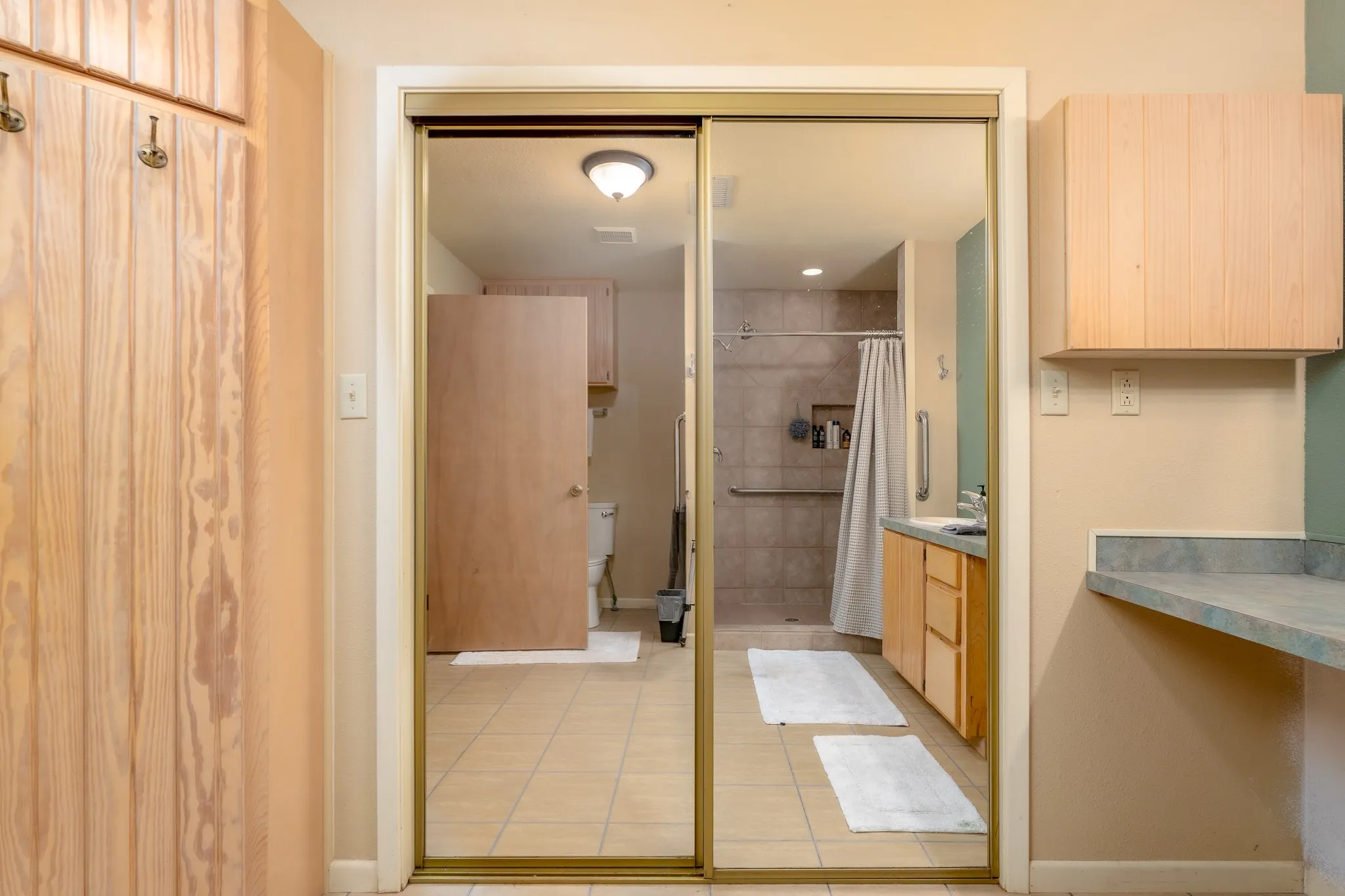 Mirrored closet doors revealing an accessible bathroom with a roll-in shower, natural wood-finish cabinetry, and tiled flooring