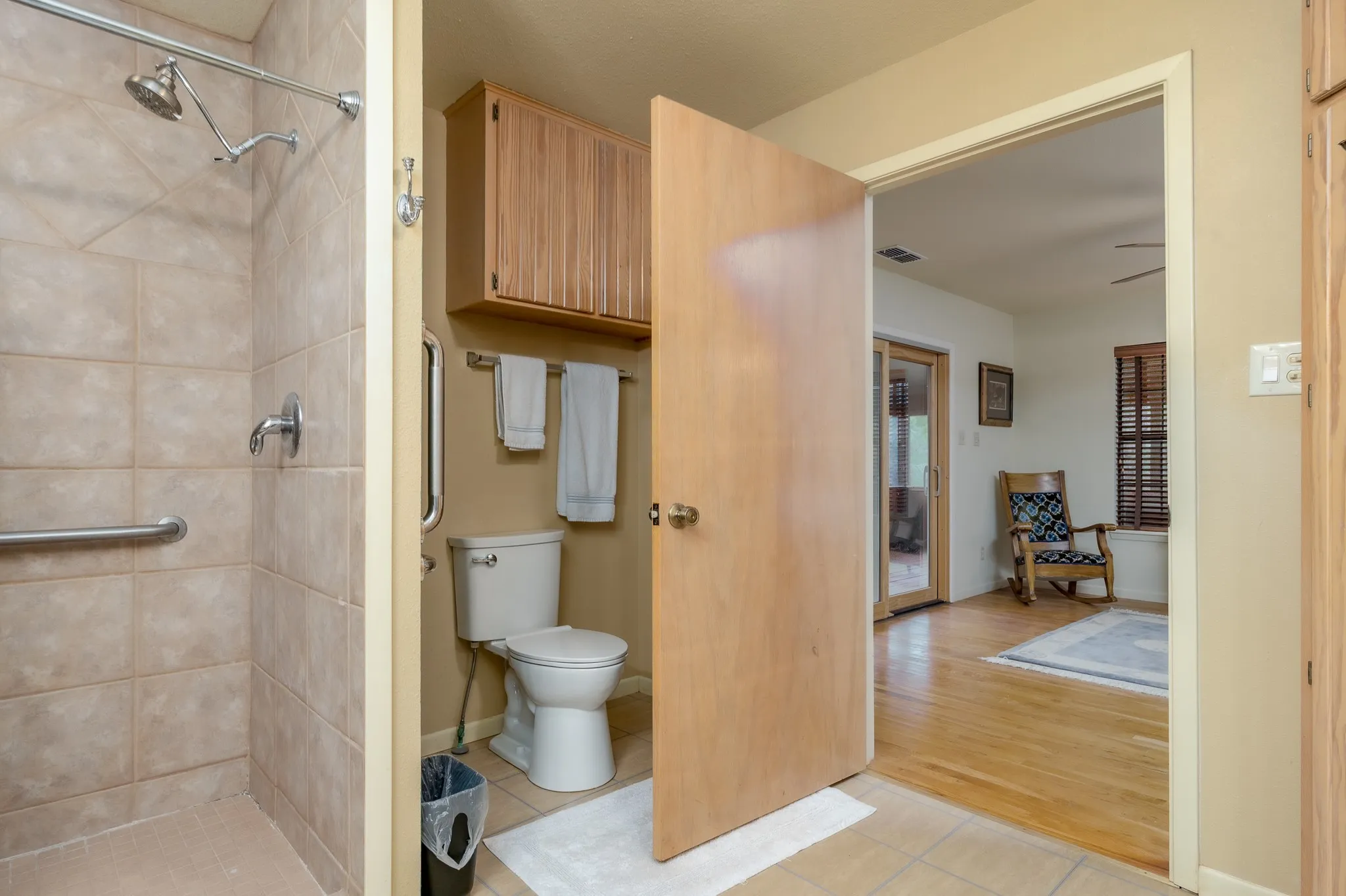 Bathroom featuring a tiled walk-in shower with grab bars and a chrome shower head, a toilet, and an overhead wood-finish cabinet