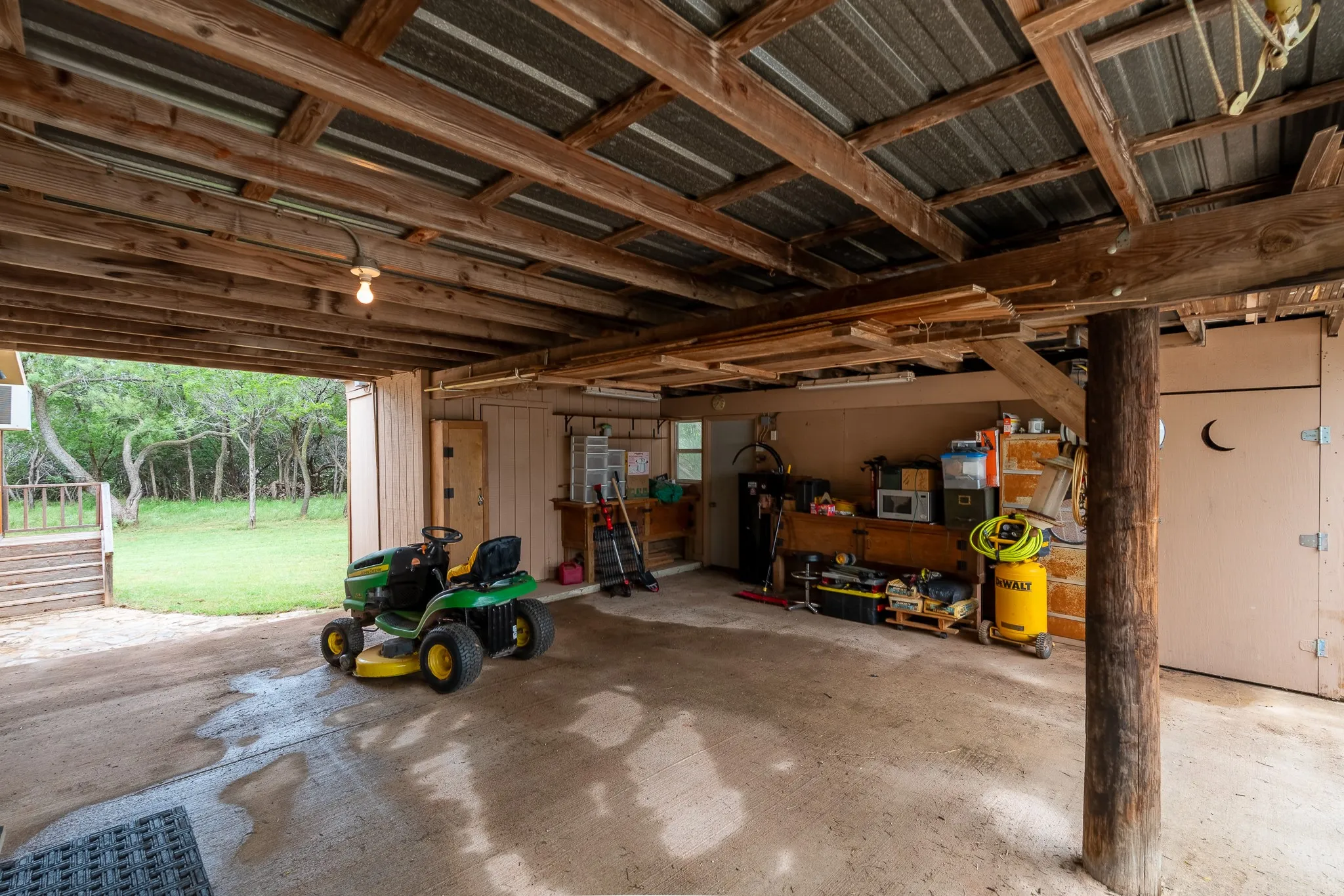 Covered concrete parking area featuring an exposed beam ceiling, corrugated metal roofing, and direct access to an enclosed storage space