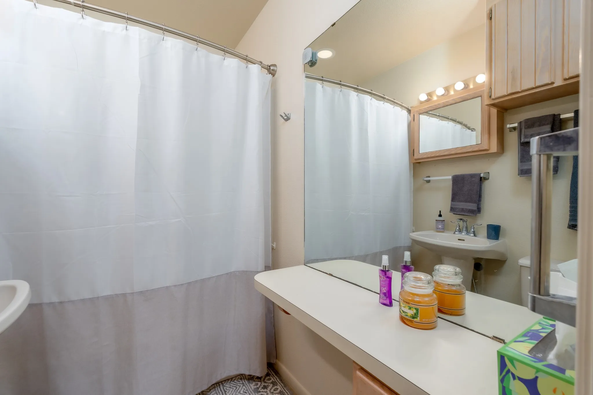 Bathroom featuring a pedestal sink with chrome faucet, light wood-finish cabinetry, vanity mirror with integrated lighting, and a full shower with a curved rod