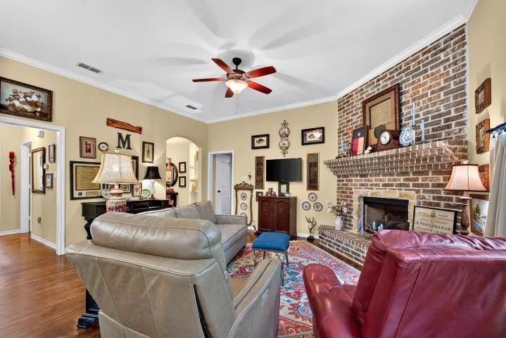 Living area featuring a brick fireplace with a raised hearth and mantel, wood-finish flooring, a ceiling fan with integrated lighting, and white crown molding