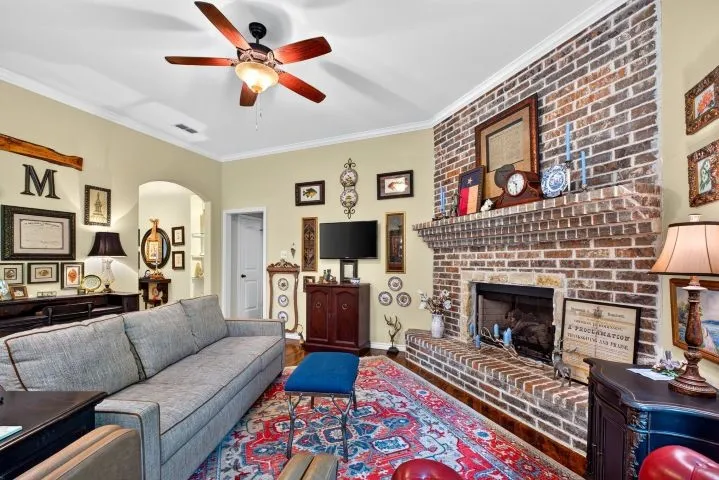 Spacious living area featuring a brick fireplace with stone hearth, crown molding, a ceiling fan with light fixture, and an arched doorway