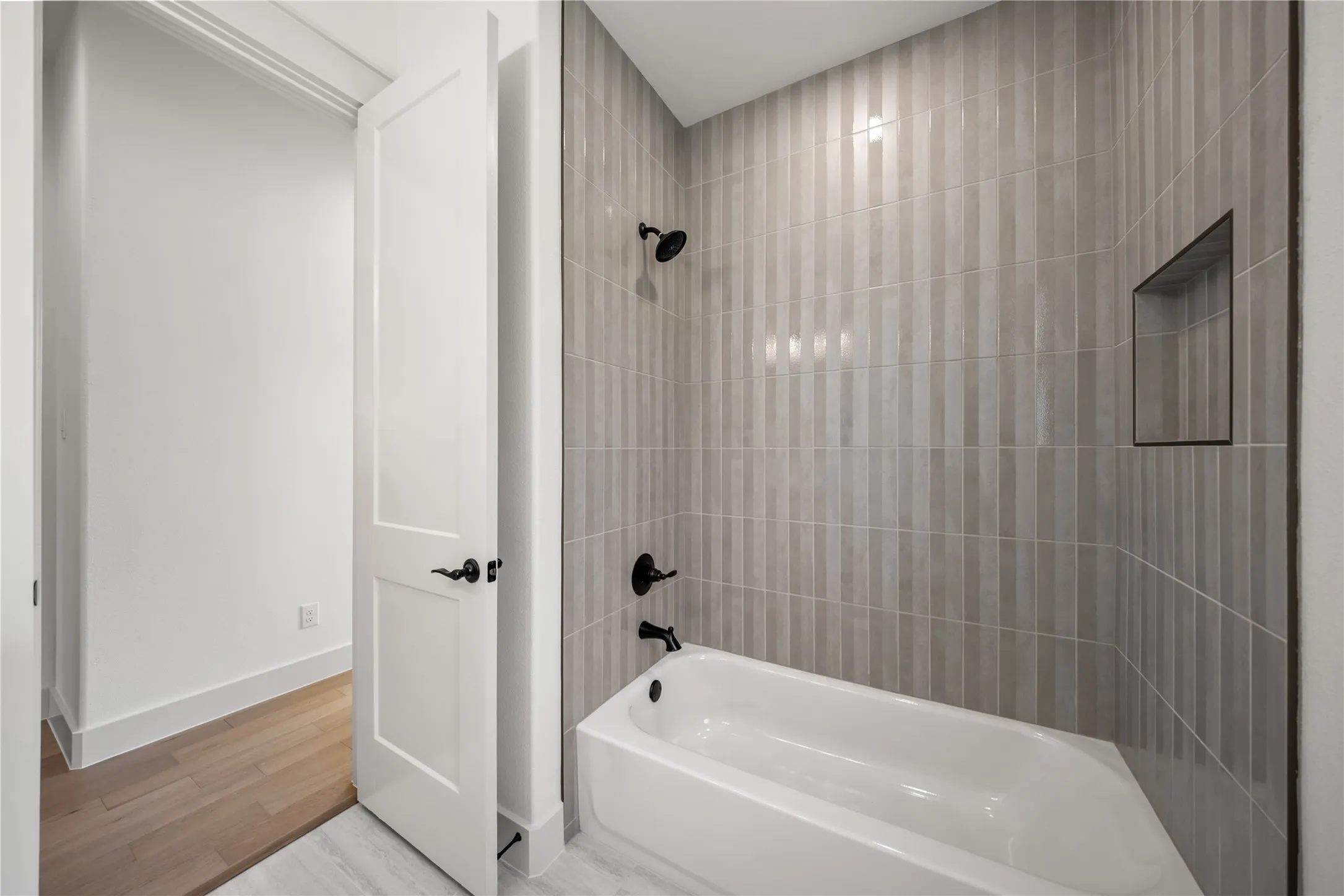 Modern bathroom featuring a white soaking tub with matte black fixtures, an integrated tiled shower niche, and vertical light gray tiled walls