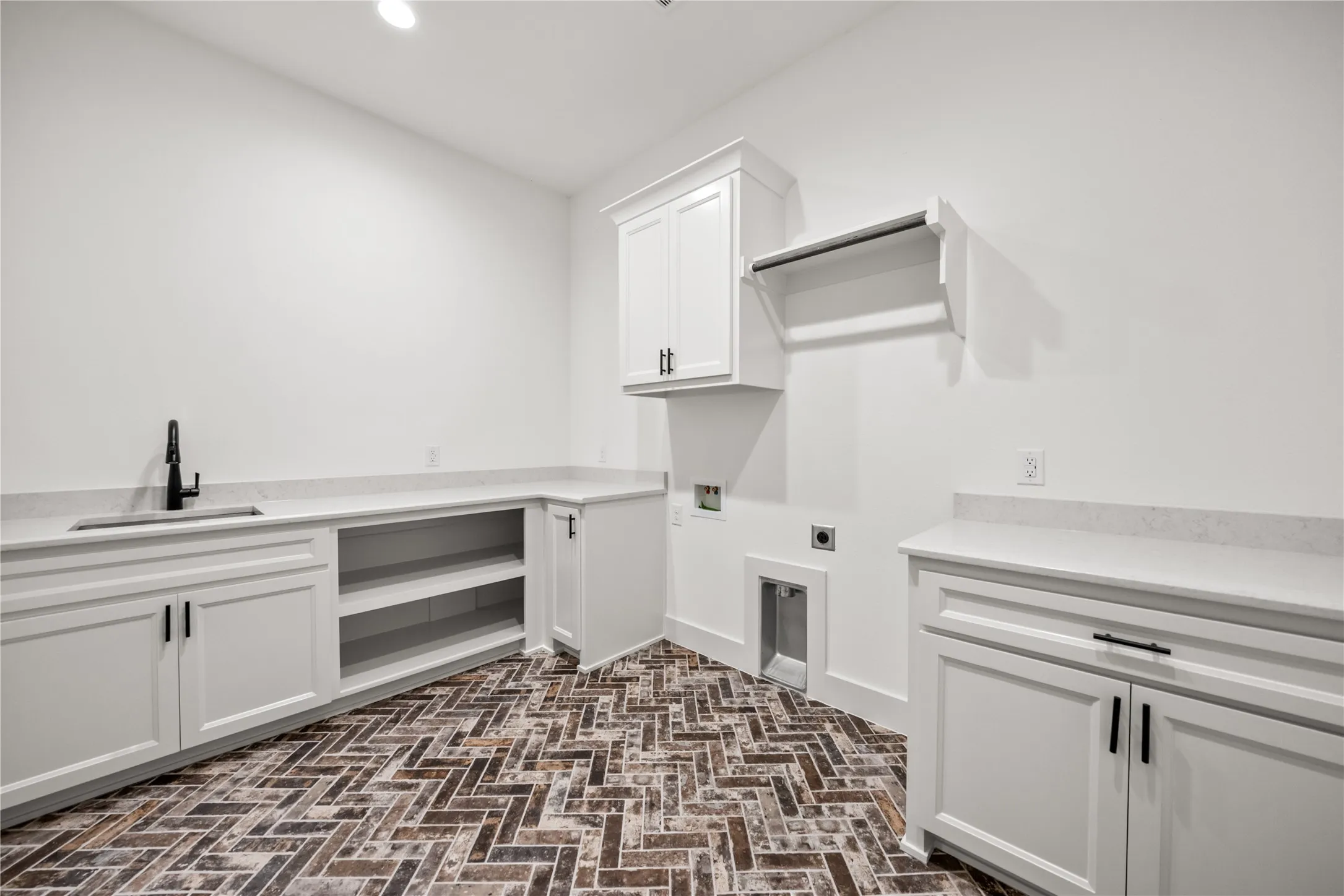 Laundry area featuring white cabinetry with black hardware, extensive counter space, an integrated sink with a black faucet, herringbone-patterned brick flooring, and built-in shelving