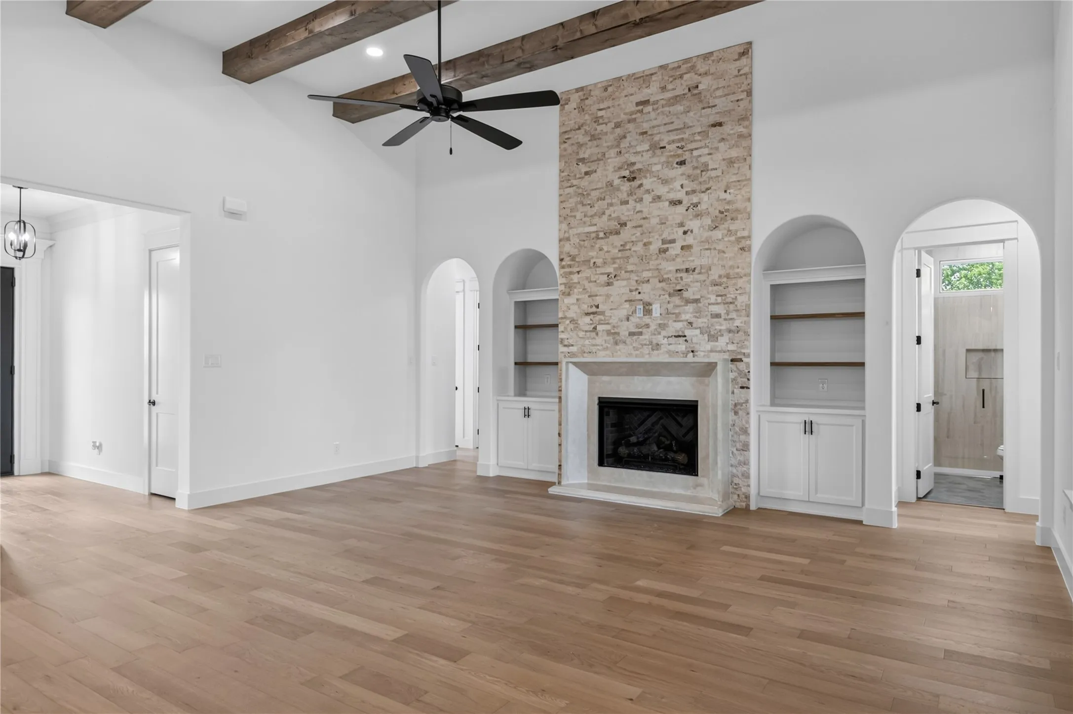 Living area featuring a stone-clad fireplace with hearth, vaulted ceiling with exposed wood beams, wood-finish flooring, and built-in arched shelving with lower cabinetry