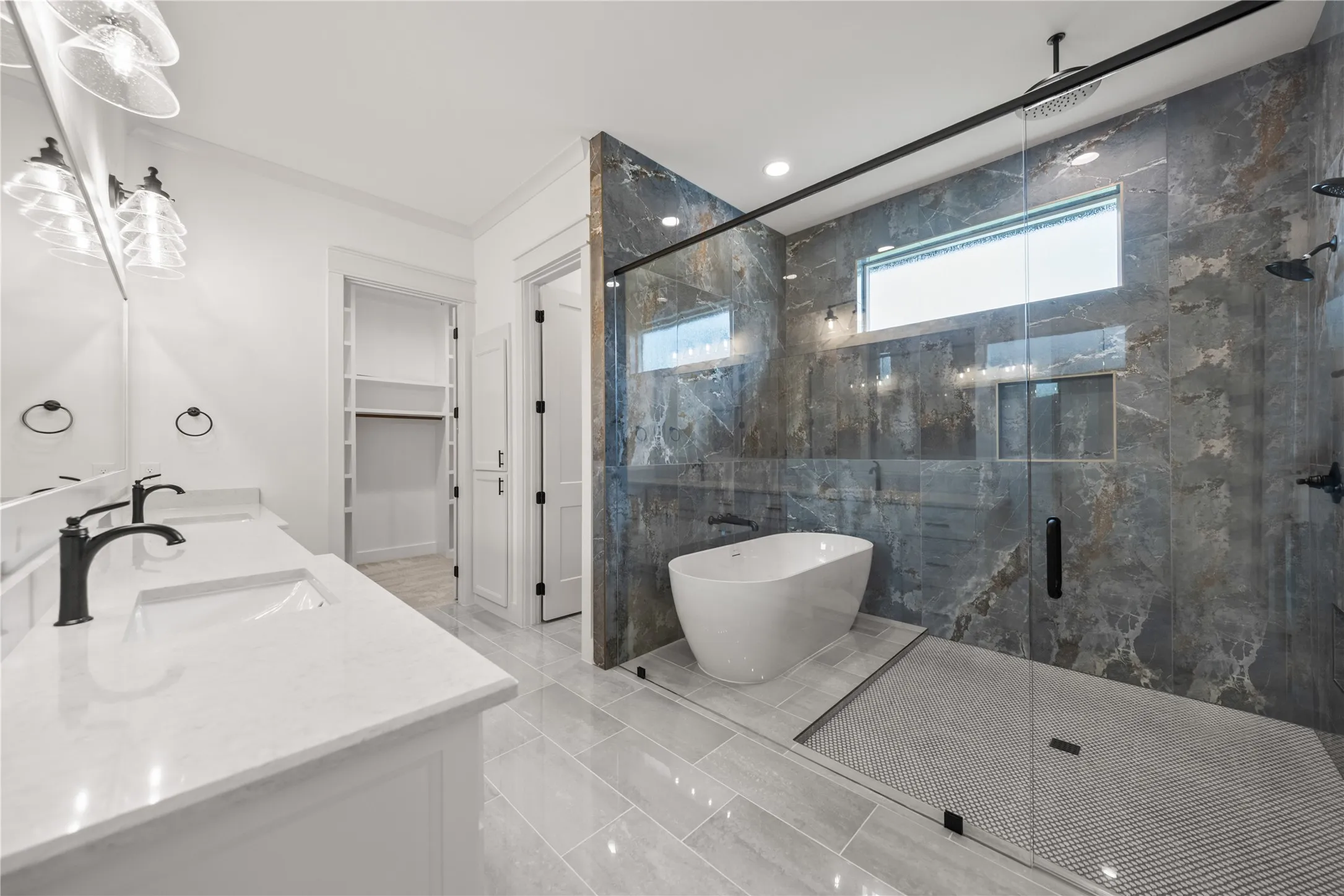 Spacious bath featuring dual vanities with white countertops, a freestanding soaking tub, and a glass-enclosed shower with dark stone-look tile