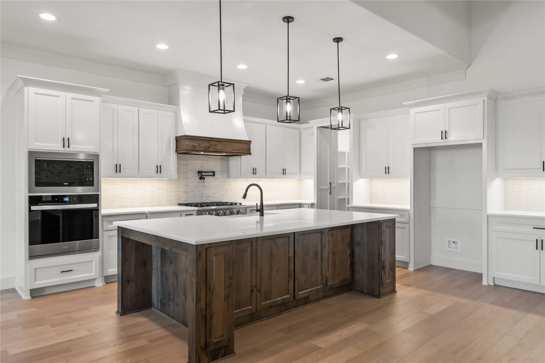 Spacious kitchen featuring a large central island with a light-toned countertop, white cabinetry with dark hardware, stainless steel wall oven and microwave, a white tile backsplash, and light wood-finish flooring