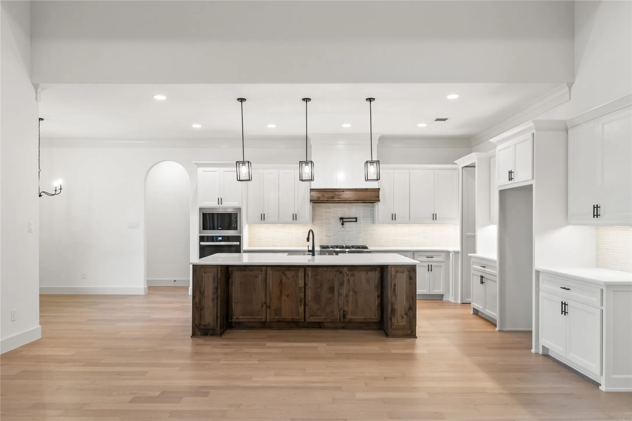 Spacious kitchen featuring a large central island with a wood-finish base and light-toned countertop, white cabinetry with dark hardware, built-in stainless steel appliances, and wood-finish flooring