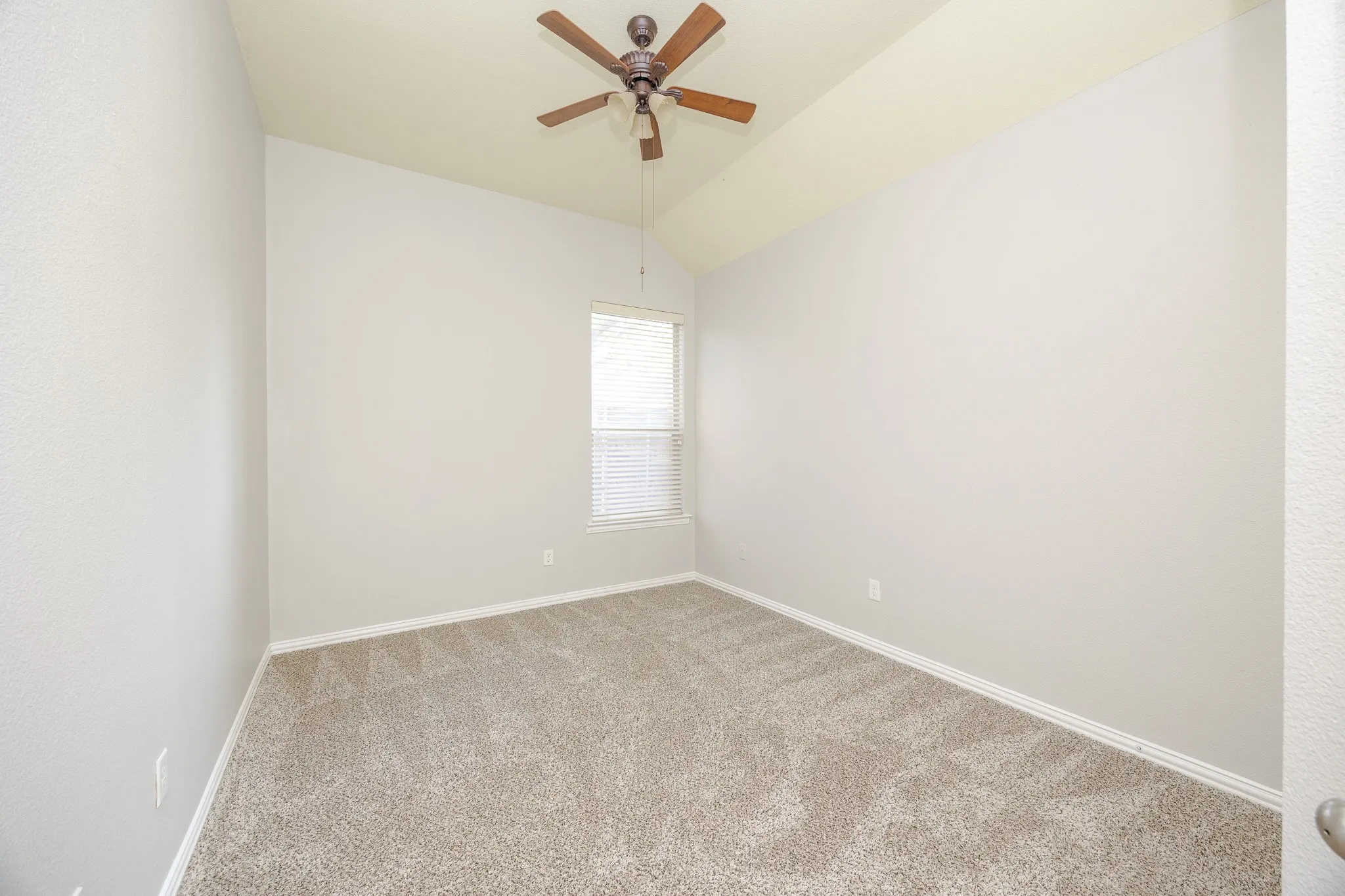 Vaulted ceiling, ceiling fan with wood-finish blades, window with blinds, light gray wall paint, and neutral carpet