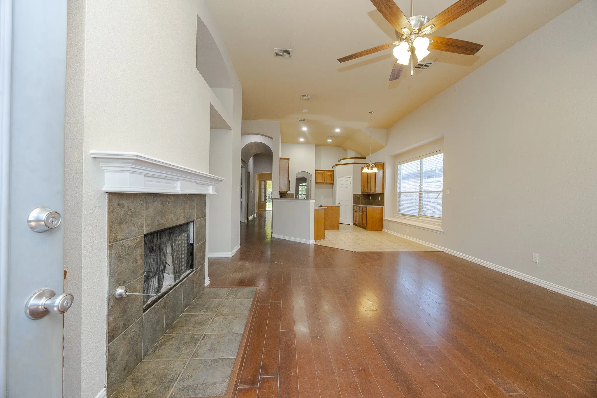 Fireplace with a tile surround and white mantel, wood-finish flooring, vaulted ceiling, ceiling fan with integrated lighting, and a large window