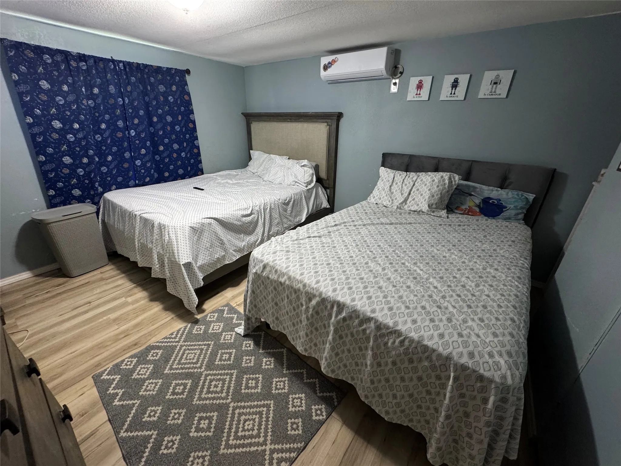Well-lit room featuring light wood-finish flooring, a wall-mounted air conditioning unit, and a window with dark blue curtains