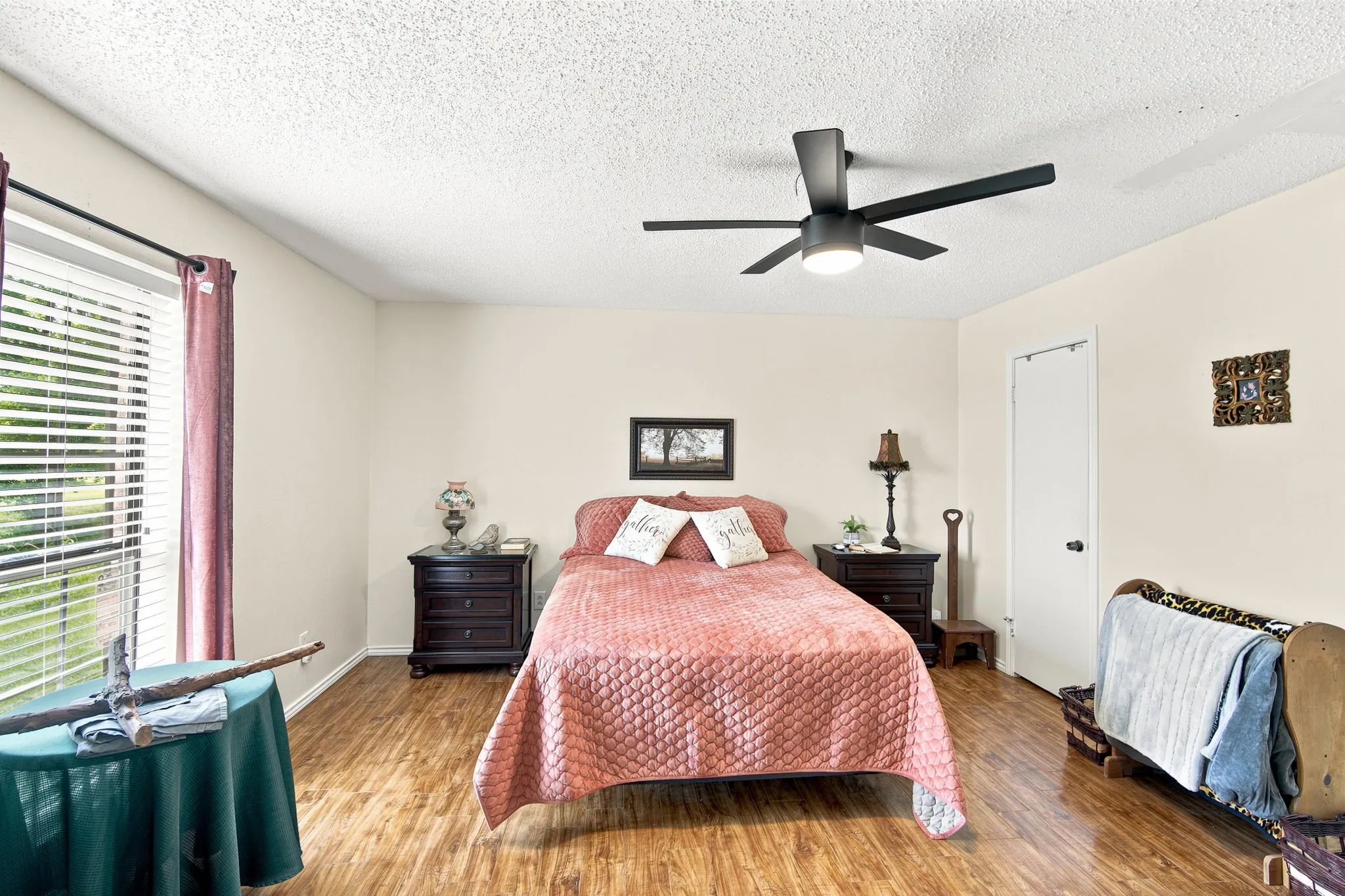 Spacious bedroom featuring wood-finish flooring, a contemporary ceiling fan with integrated lighting, and a window with horizontal blinds